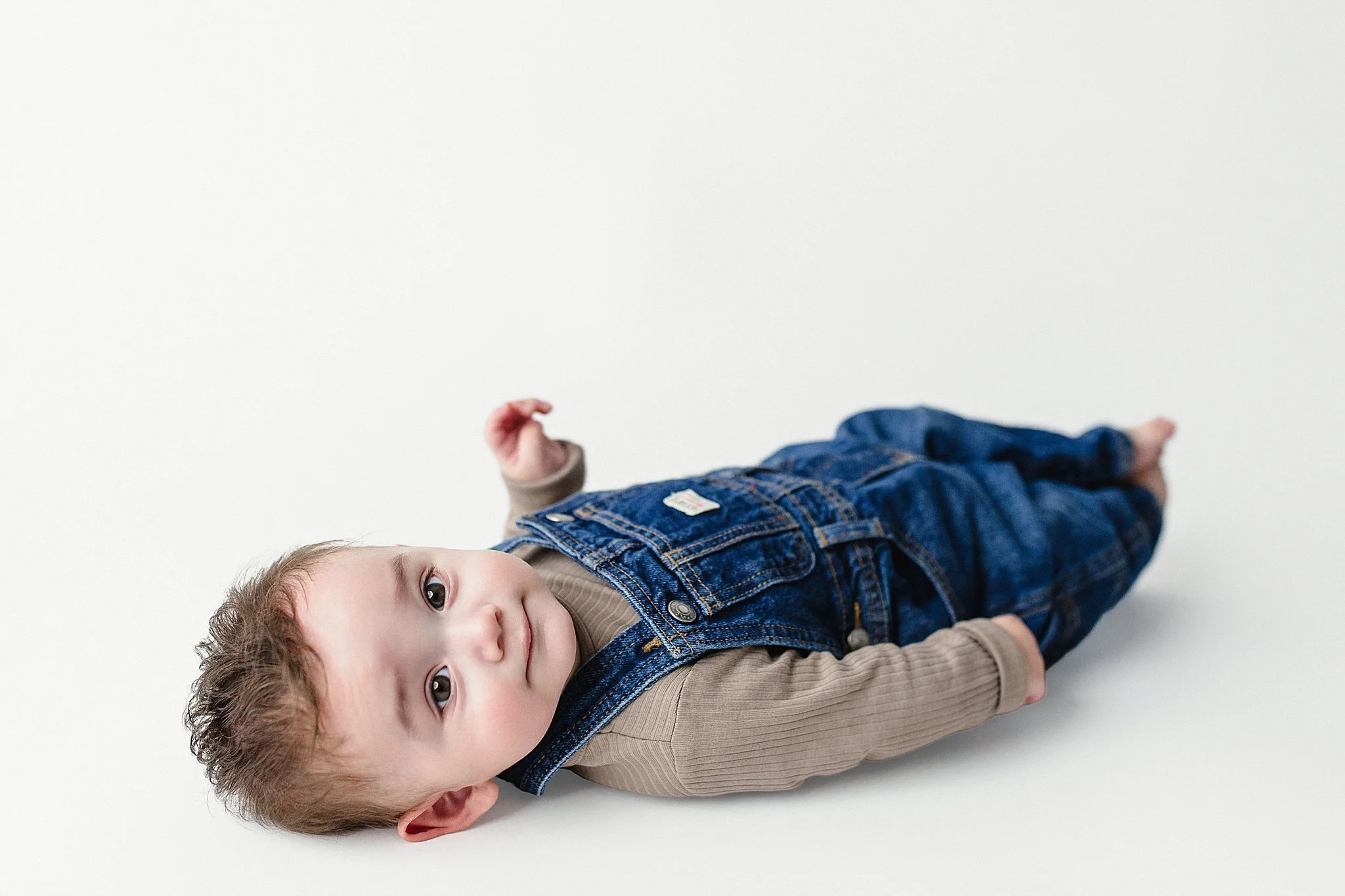 Young child lying on a white surface, looking at the camera, wearing a tan long-sleeve shirt and blue denim overalls.