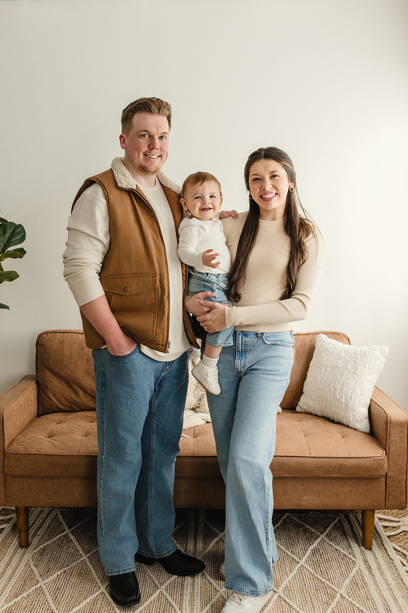 A family of three standing in a living room, smiling at the camera. The father is wearing a brown vest and jeans, the mother is dressed in a beige sweater and jeans, and the young child is held by the mother during a lifestyle photoshoot.
