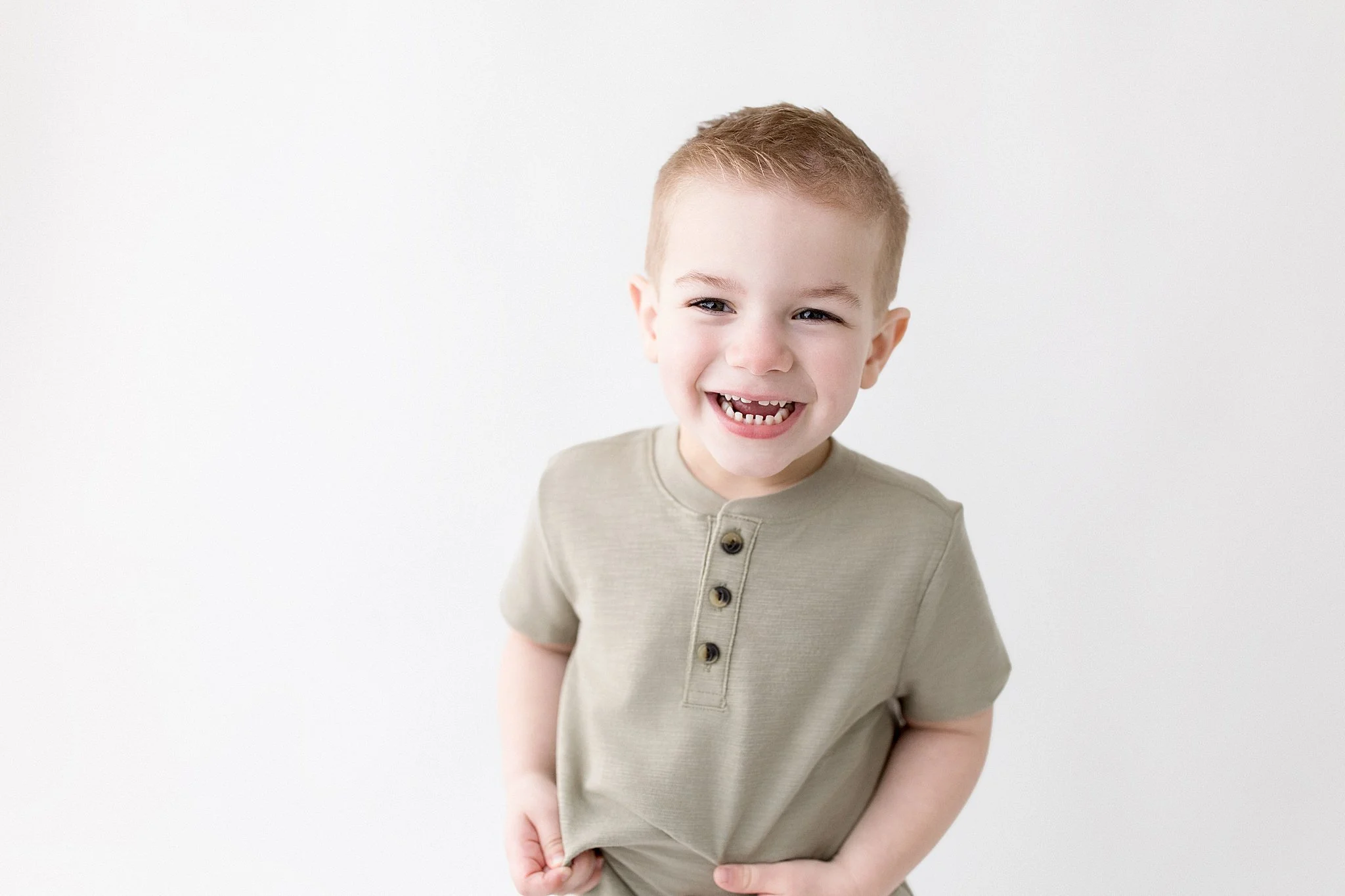 A young boy with short light brown hair smiling, showing missing front teeth, wearing a beige t-shirt with a button placket, standing against a plain white background for kids acting photos near me. 