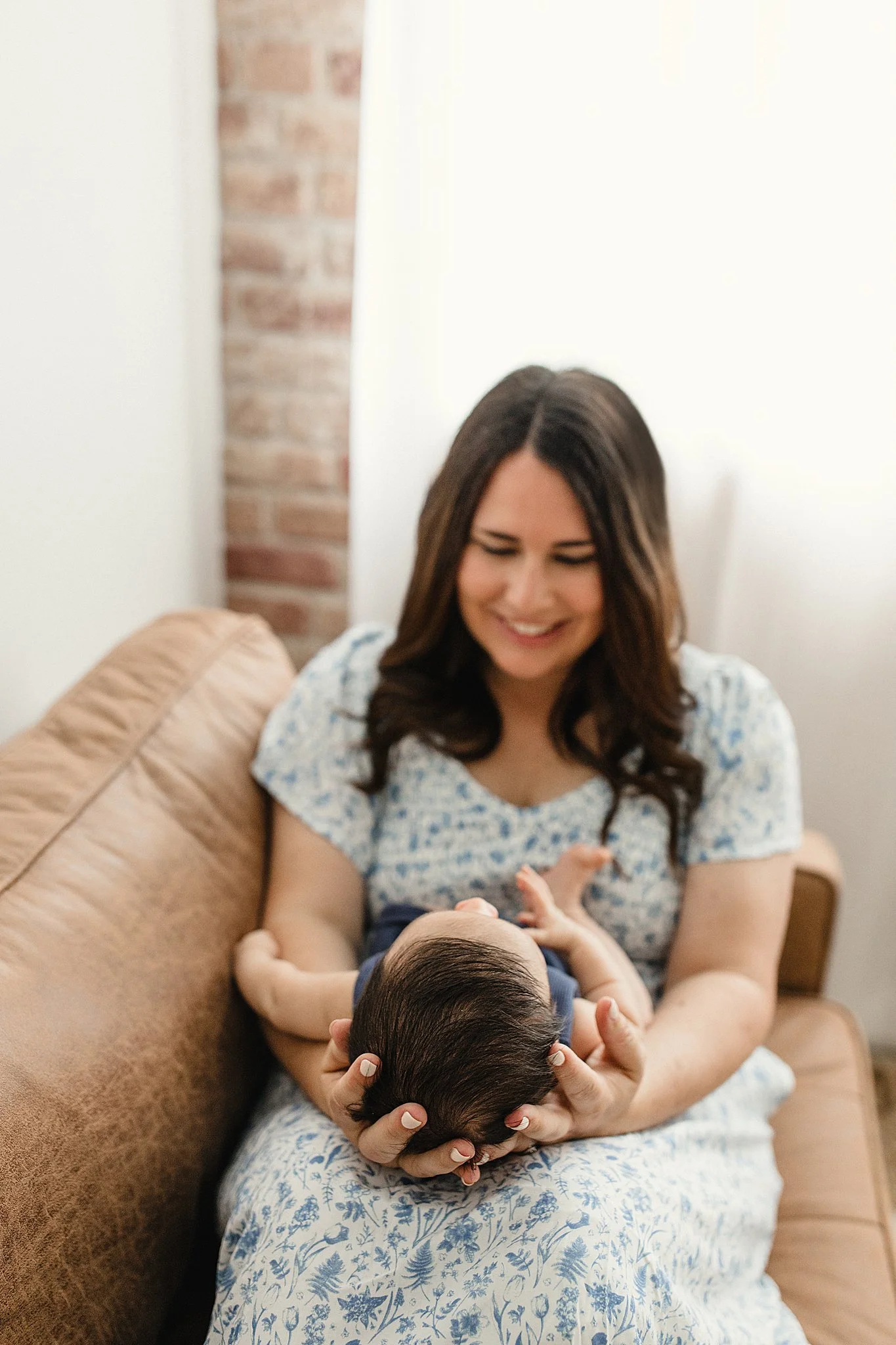 A smiling woman sitting on a brown couch, holding a small baby in her lap. The woman has long dark hair and is wearing a light blue and white floral dress during a newborn photo session in Naperville, IL.