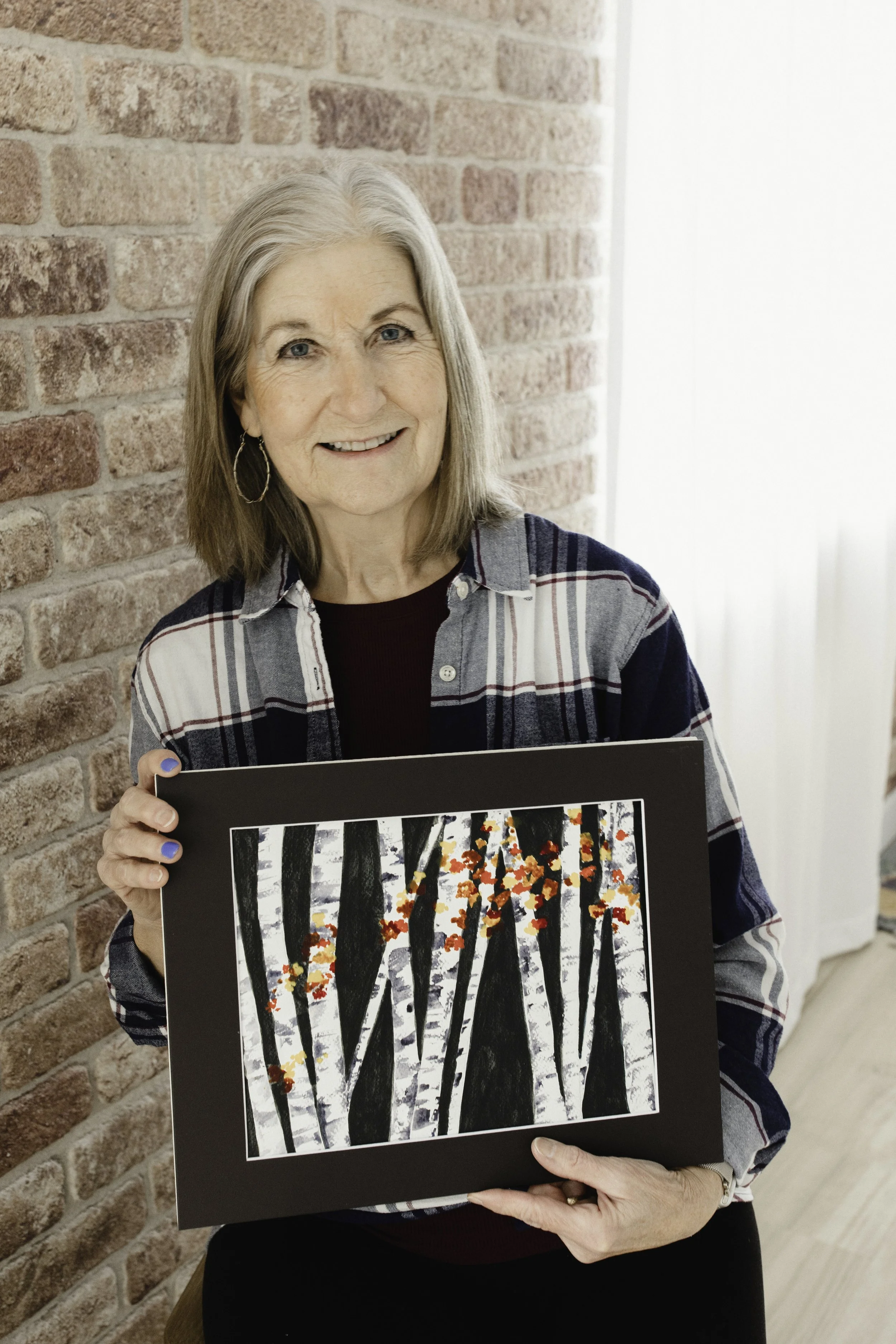 An elderly woman with shoulder-length gray hair, wearing a plaid shirt, holding a framed painting of a forest of white birch trees with red and orange leaves, during a watercolor painting class taught by Jonnette Maslowski in Naperville, IL.