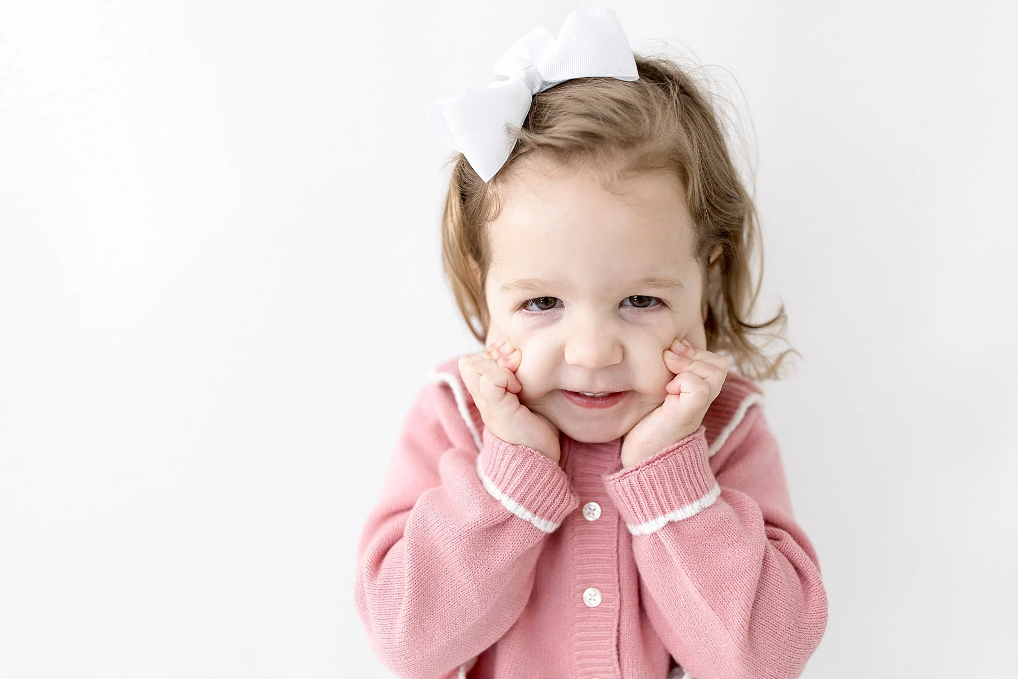 A young girl with a white bow in her hair, wearing a pink sweater, pressing her cheeks with her hands, and smiling at the camera against a white background during a personality mini session near me.