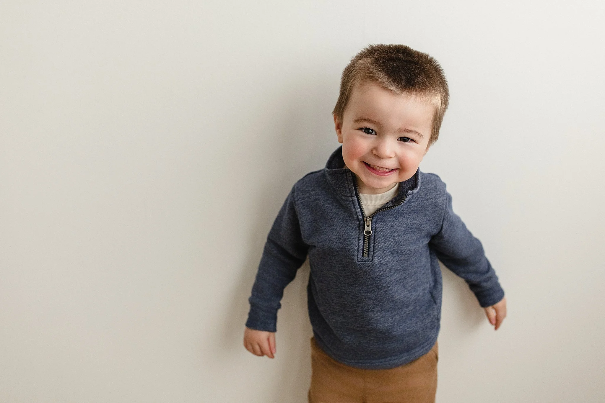 Smiling young boy with short brown hair wearing a navy blue quarter-zip sweater and tan pants standing against a plain off-white wall with a family photographer near me.