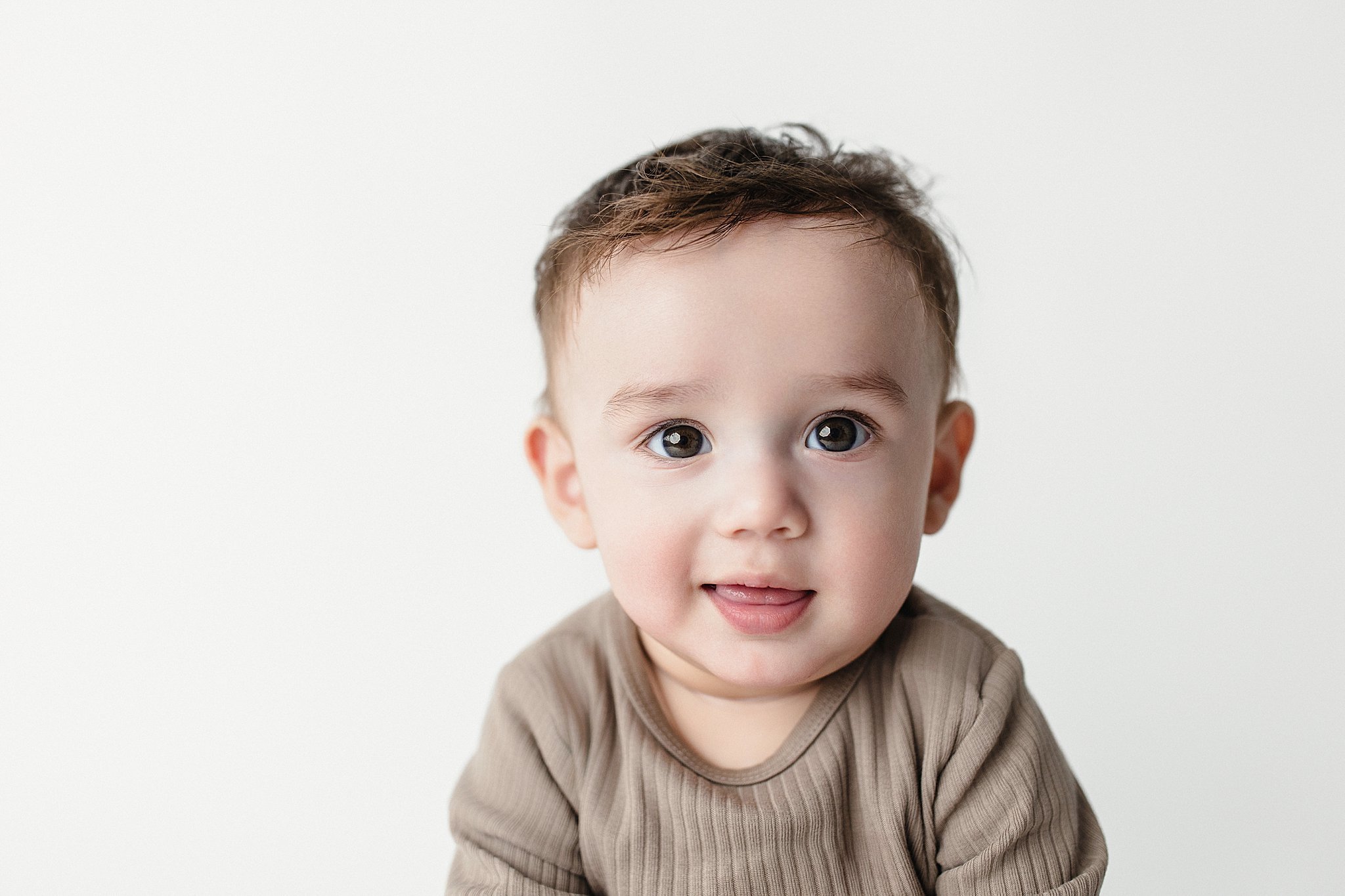 Close-up of a smiling young child with short brown hair and big brown eyes, wearing a beige shirt, against a plain white background during a baby photoshoot Naperville, IL.