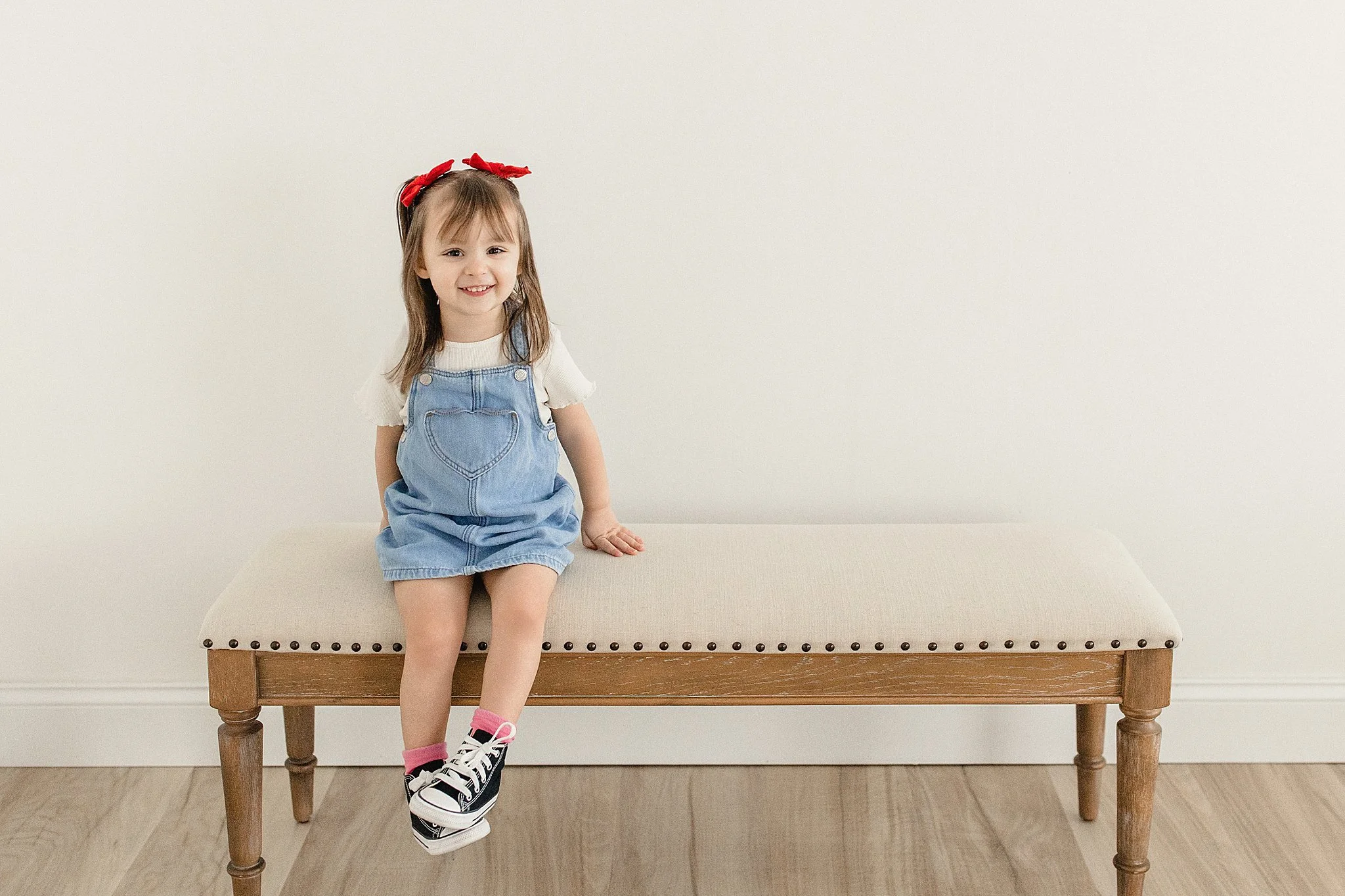 A young girl sitting on a beige upholstered bench against a plain white wall. She is smiling, wearing a white t-shirt, blue denim dungarees with a heart-shaped pocket, black and white sneakers, and pink socks. She has a red bow headband in her brown 