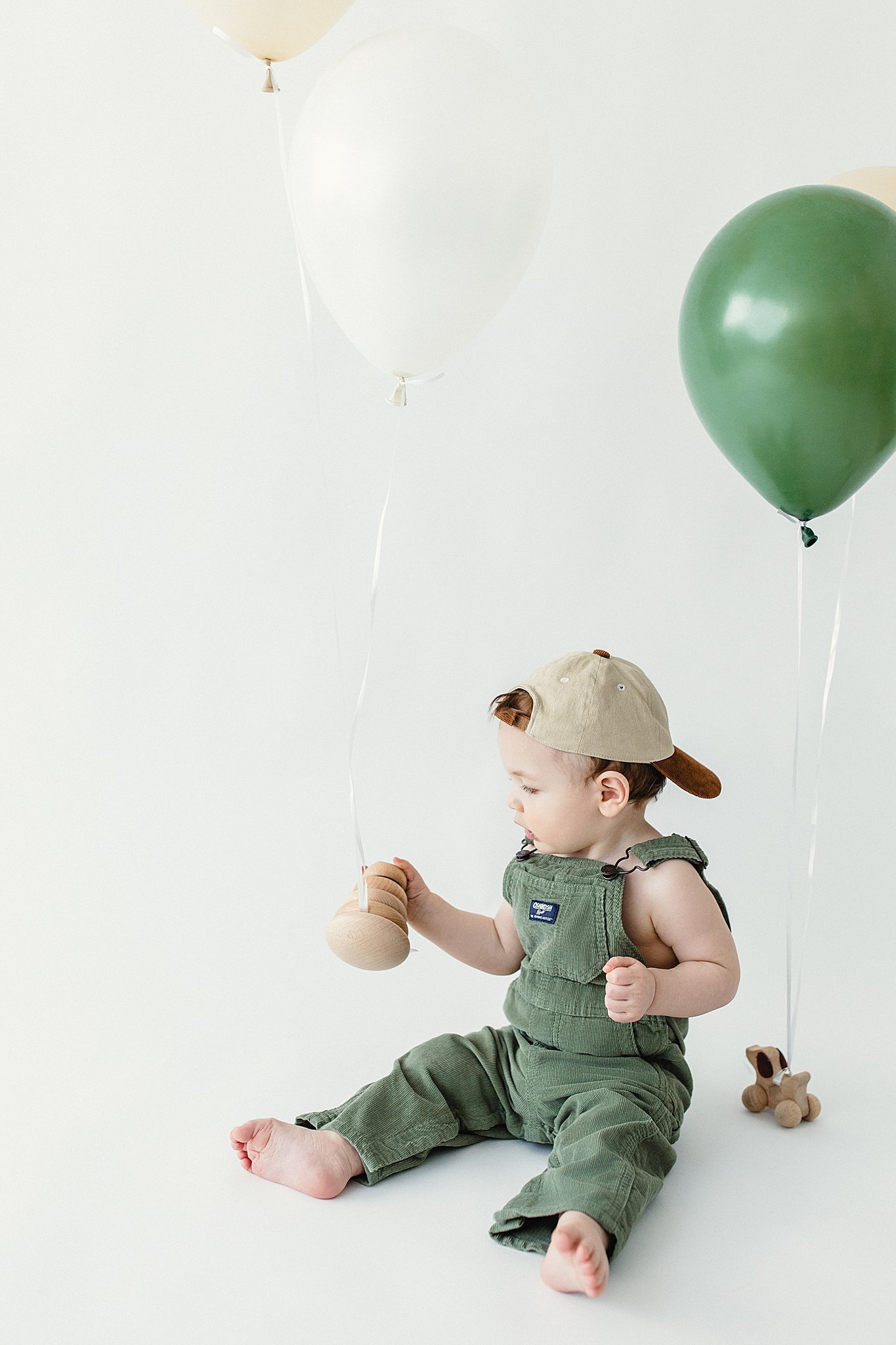 A baby sitting on the floor holding a wooden rattle in one hand, wearing green overalls and a beige cap, surrounded by white and green balloons during first birthday photos in Naperville, IL.