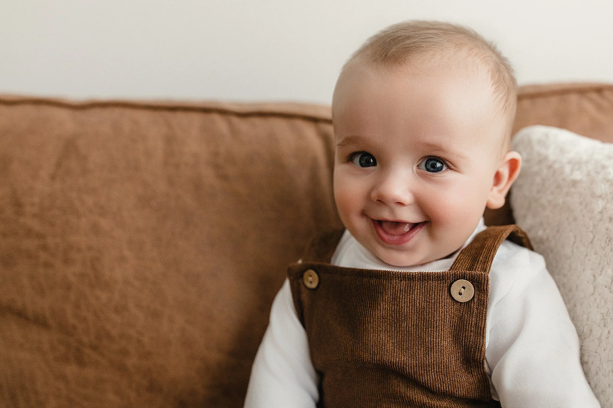 A smiling baby with blue eyes and light hair sitting on a brown couch, wearing a white shirt and brown overalls for a baby photoshoot in Naperville, IL.