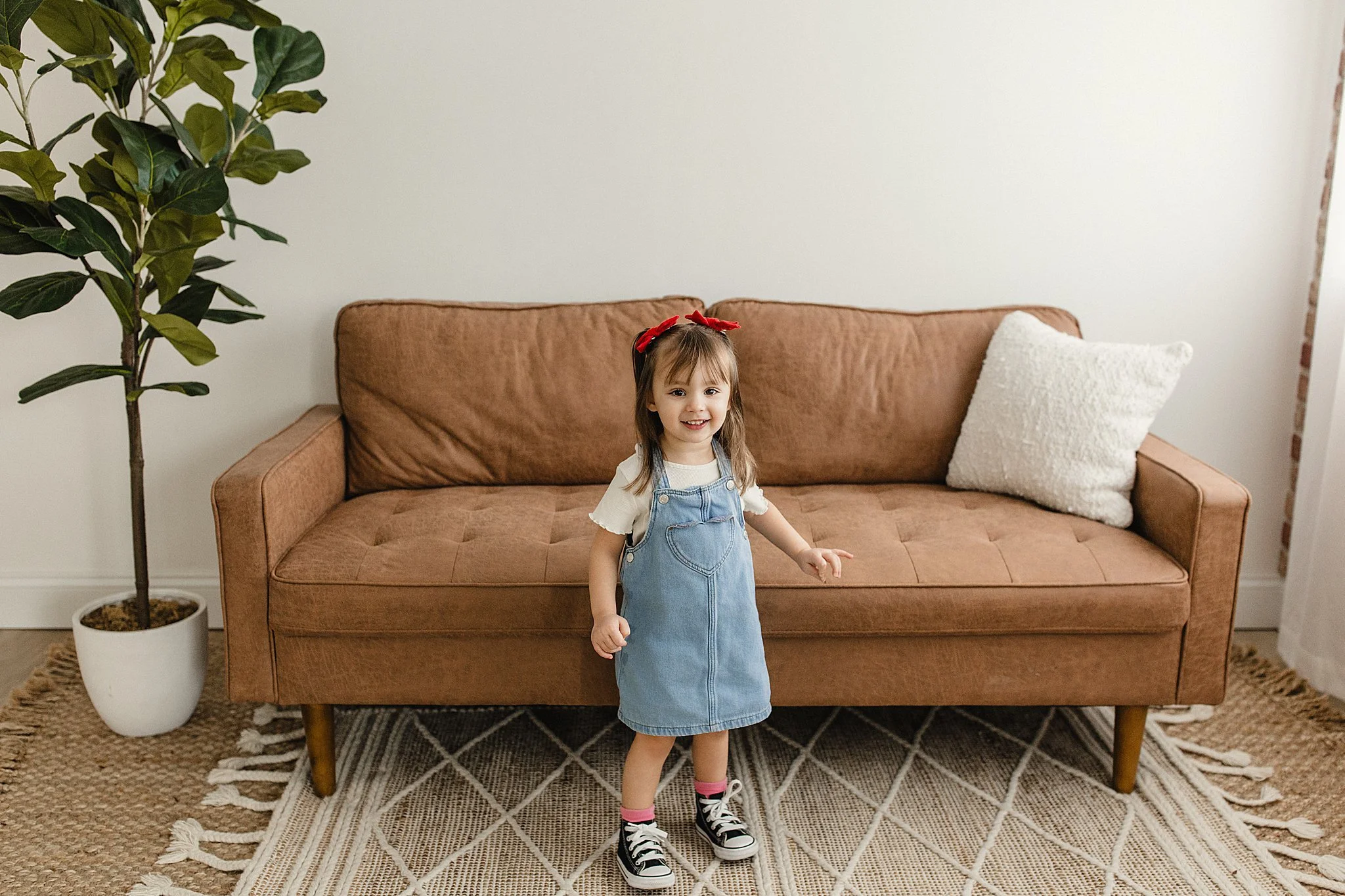 A young girl with a red bow in her hair, wearing a white t-shirt, denim jumper dress, and black sneakers with pink socks, standing in front of a brown sofa with a white pillow, indoors next to a green potted plant, on a patterned rug during photos.