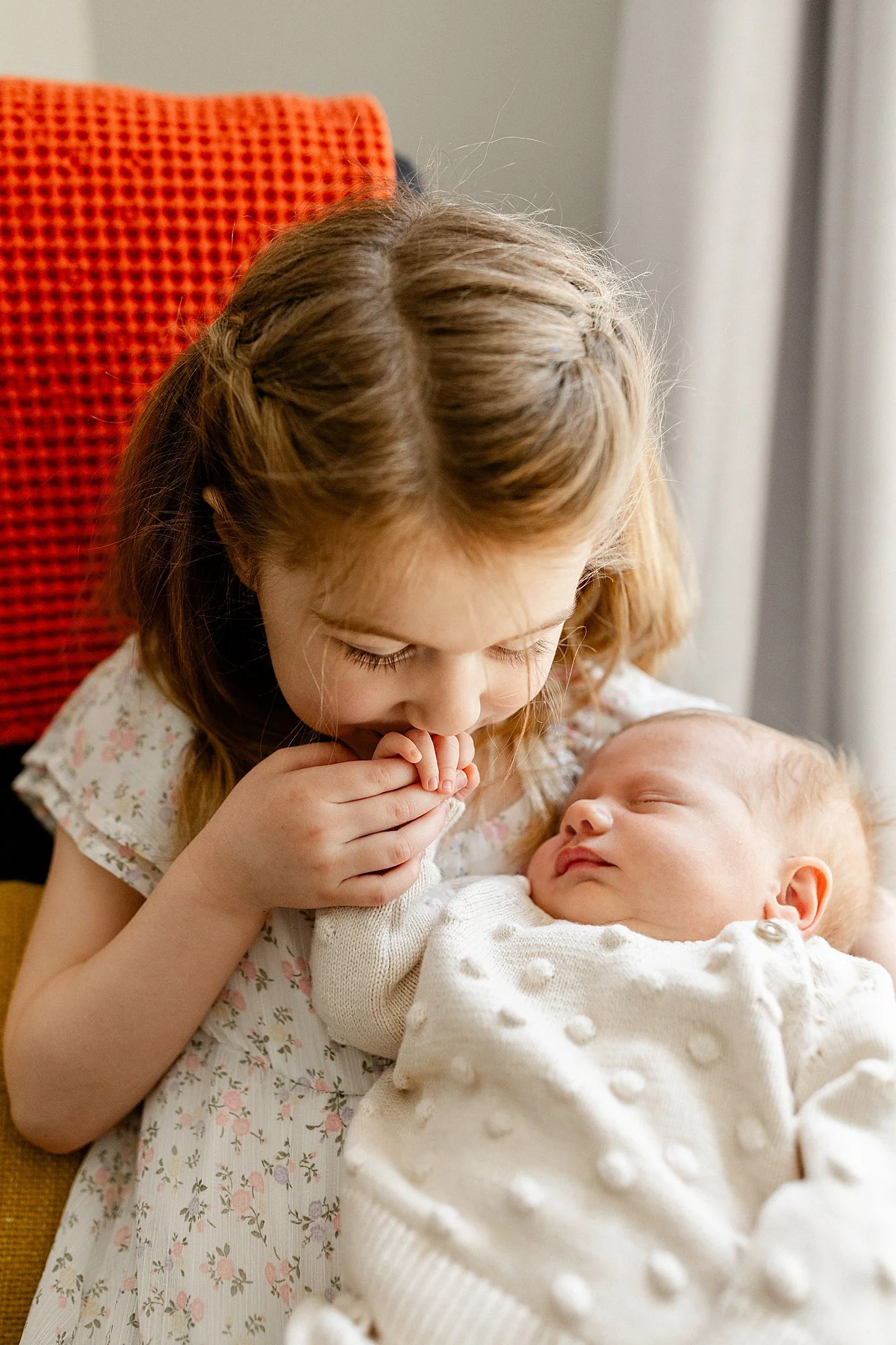 A young girl with braids looks at a sleeping baby she is holding in her arms, in a room with curtains and an orange chair.