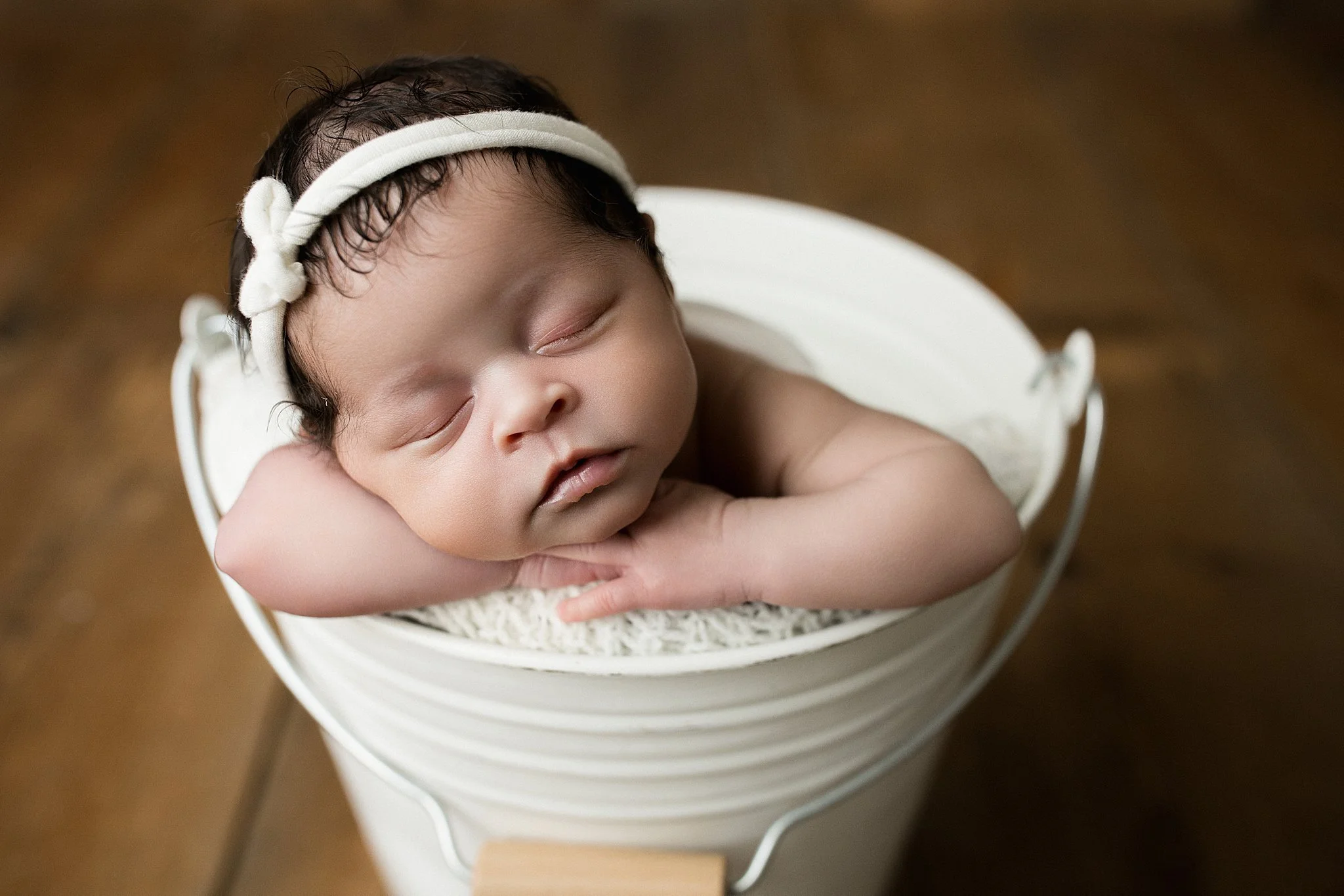 Close-up of a sleeping newborn baby with dark hair, resting with head on arms, in a white bucket with a brown background during a newborn session in Naperville, IL.