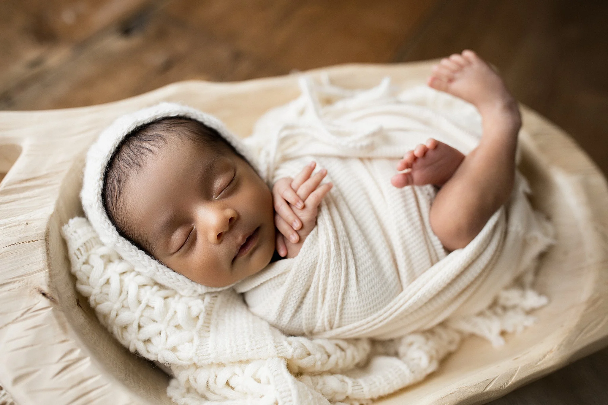 A sleeping baby dressed in a cream knit hoodie and outfit, resting on a wooden surface with a cream blanket during a posed newborn photoshoot with ally and b photography.