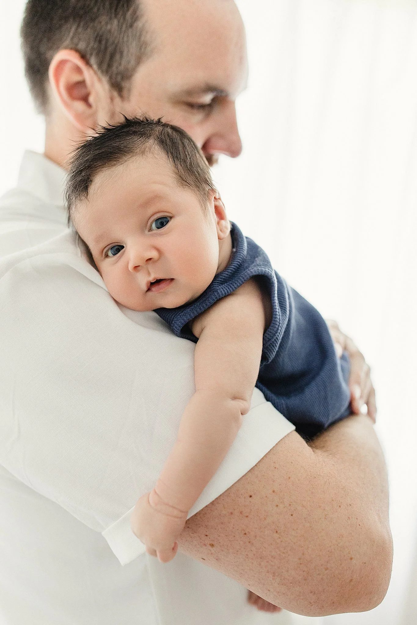 A person holding a sleeping baby on their shoulder. The baby has blue eyes and dark hair, wearing a navy blue shirt. The person's skin is fair and they are wearing a white shirt with the best newborn photographer Naperville.