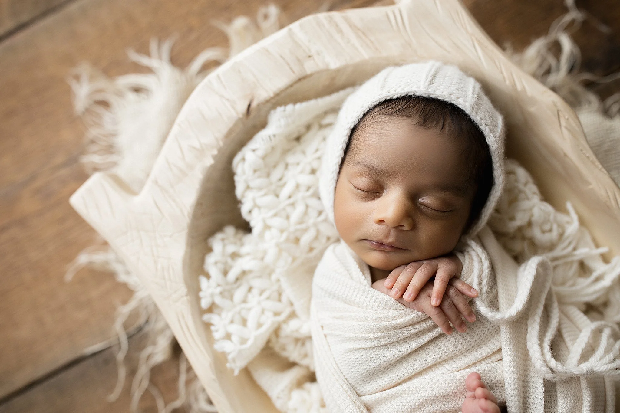 A sleeping baby wrapped in a cream-colored blanket, wearing a knit hat, lying on a wooden surface surrounded by a fringed cream-colored blanket with Ally and B Photography Naperville.