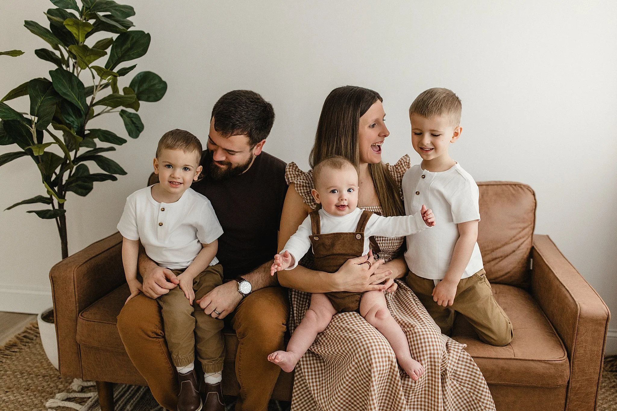 Family of five sitting on a brown sofa in a living room, smiling and interacting with each other. There are two young boys, a woman, a man, and a baby girl, with a large green potted plant nearby during lifestyle photography near me.