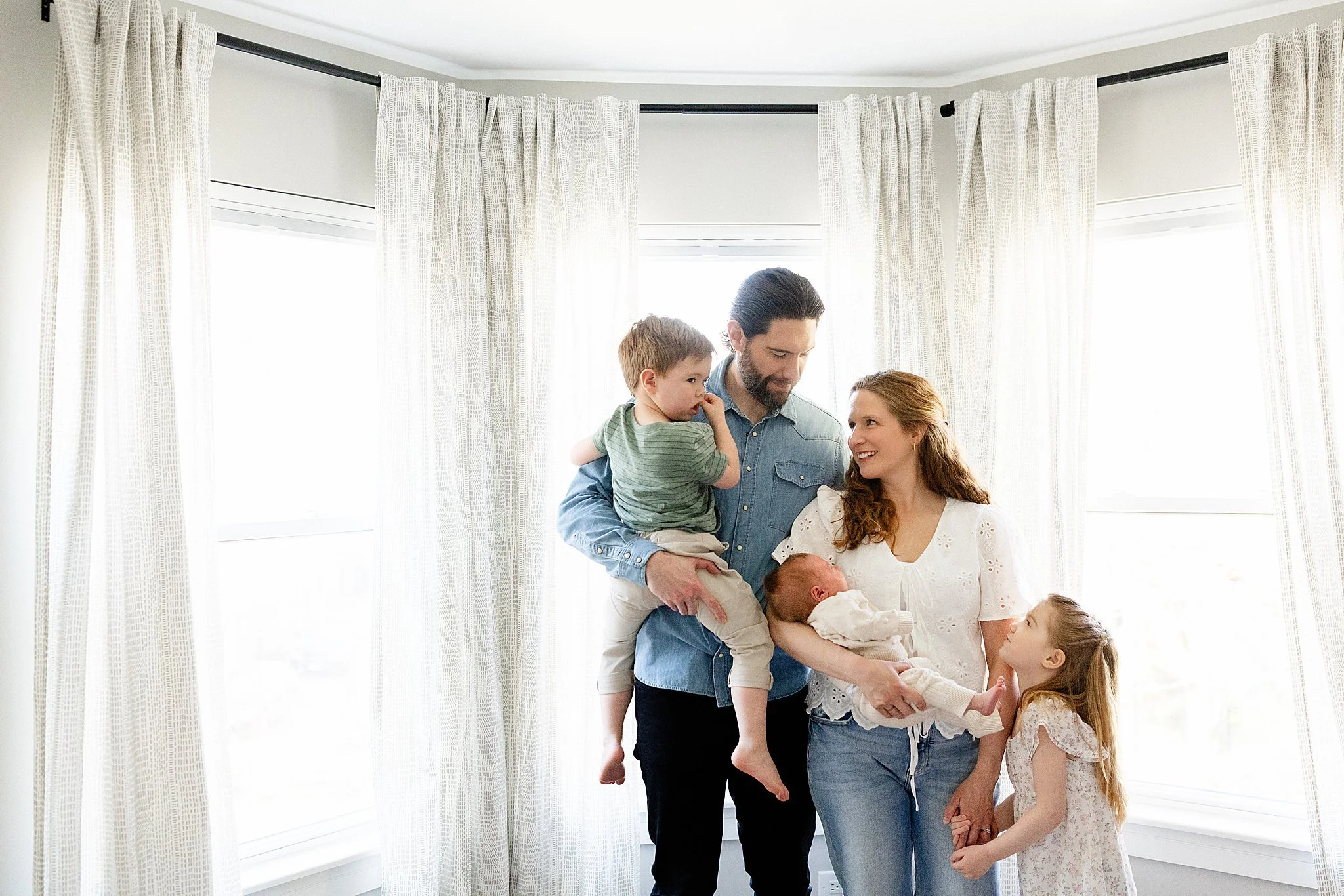 A family of seven standing in a living room near large windows with white curtains, engaging in conversation and smiling.