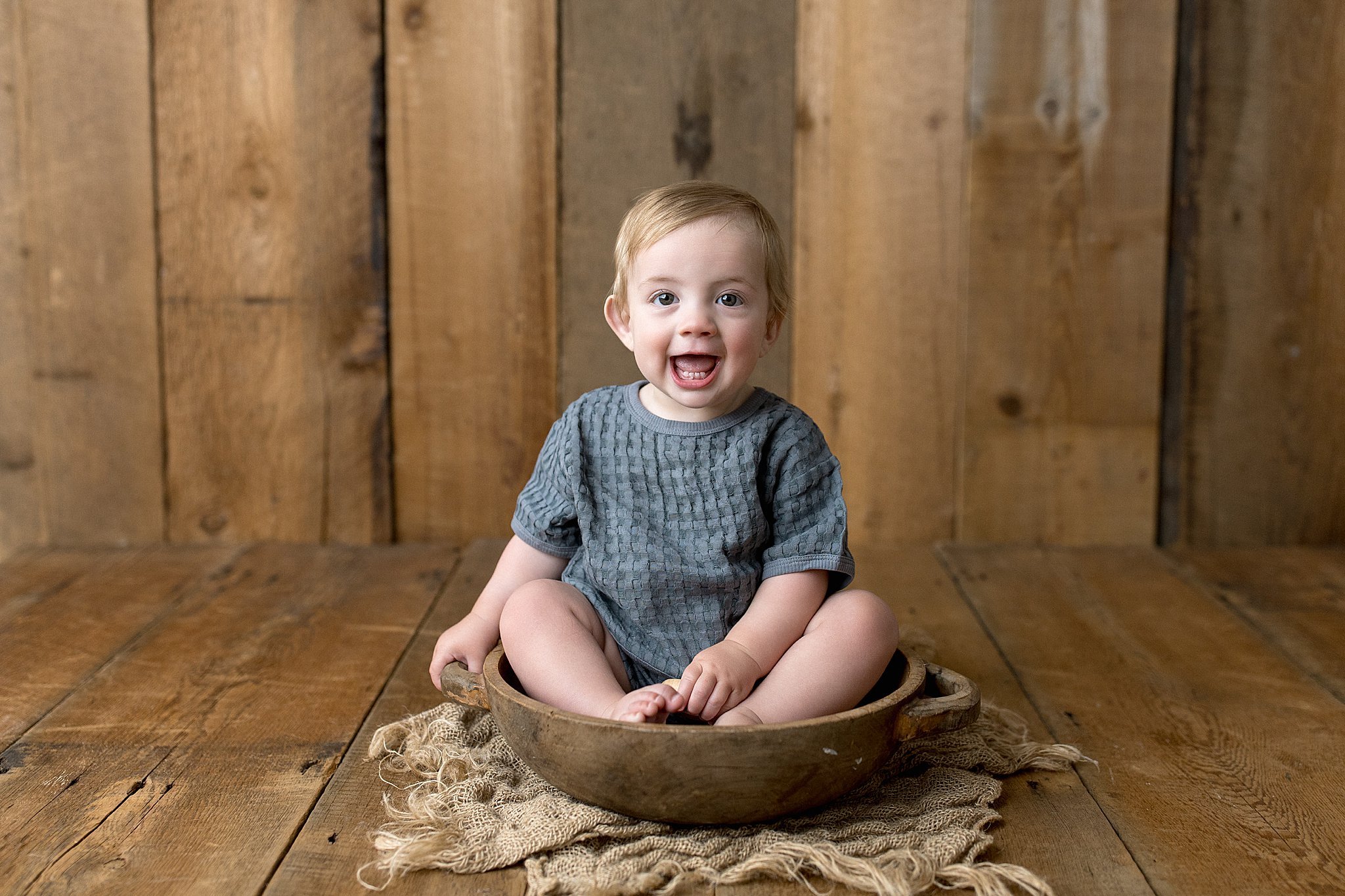 A young child with light hair and blue eyes sitting inside a wooden bowl, wearing a gray shirt, with a rustic wooden wall background with the best baby photographers near me.