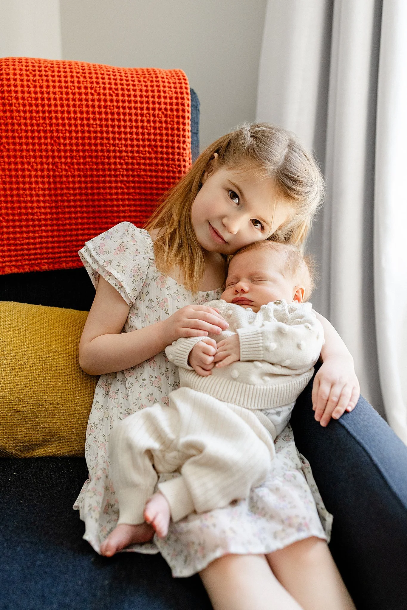 A young girl with light brown hair and a floral dress holding and cuddling a sleeping baby in a cream sweater, sitting on a dark-colored couch with colorful cushions with a lifestyle photographer Wheaton.