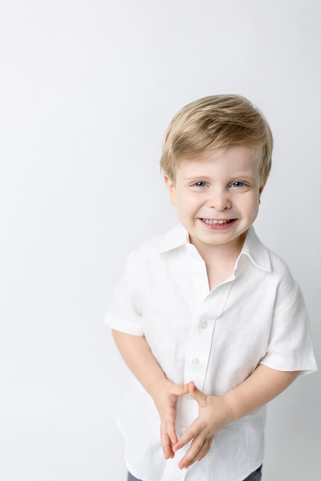 A young boy with blonde hair and blue eyes wearing a white shirt, smiling, with his hands clasped in front of him against a plain white background during a kids photoshoot in downtown Naperville, IL.