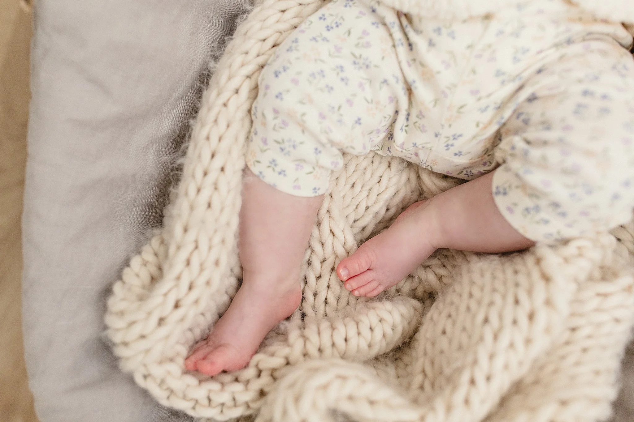 Close-up of a baby lying on a soft surface, wrapped in a chunky knit blanket, wearing a light-colored floral outfit during a newborn photography session with Ally and B Photography in Naperville, IL.