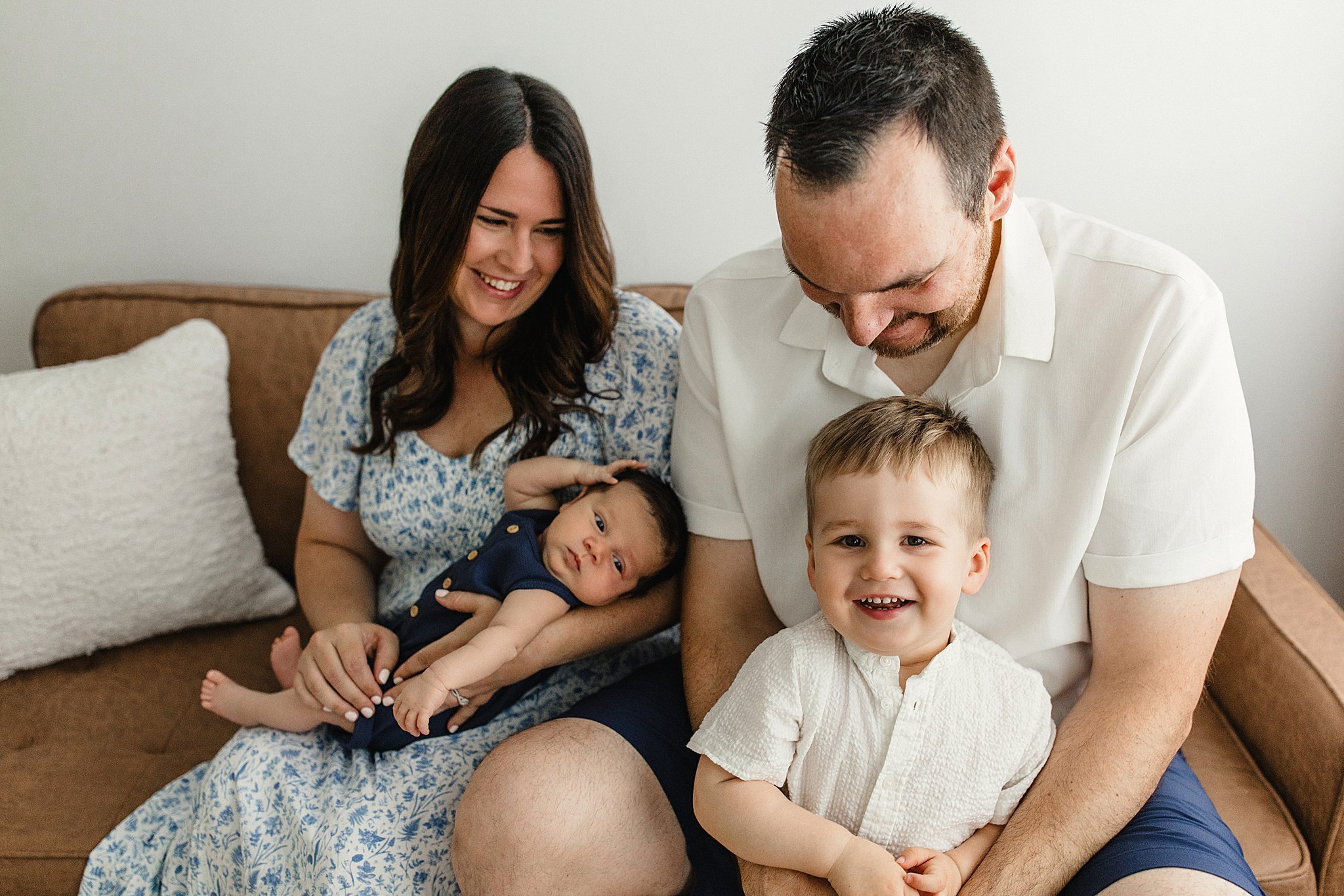 A happy family of four sitting on a brown couch. The mother with dark hair is smiling and holding a sleeping baby in her lap. The father with dark hair and beard is leaning over a young boy with light-colored hair for newborn photos near me.