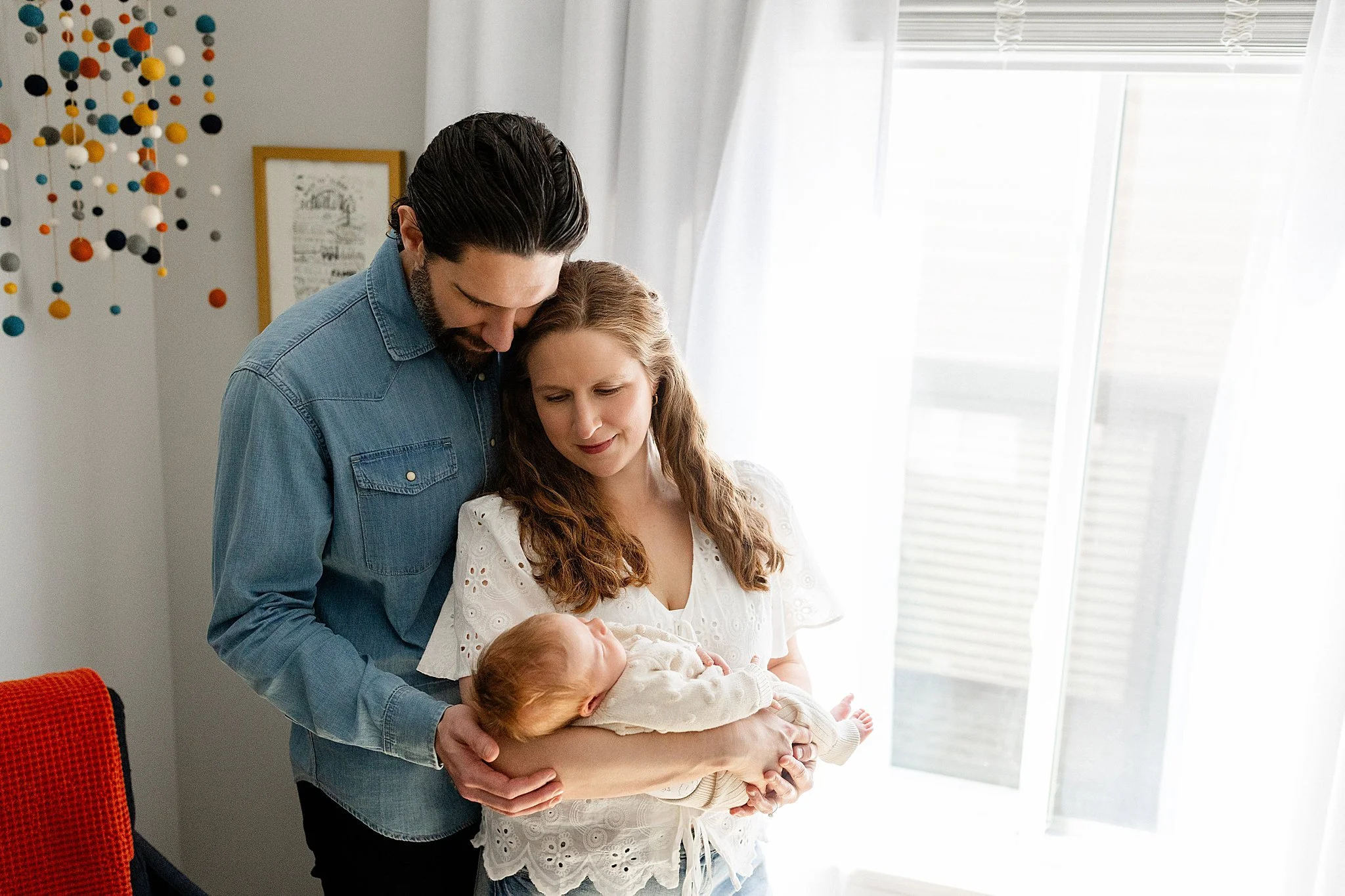 A family gathering in a bright room with curtains, including a man, a woman, and two babies. The man is leaning over and kissing a girl, the woman is holding a baby, and they are all engaged in a tender moment during lifestyle newborn photos.