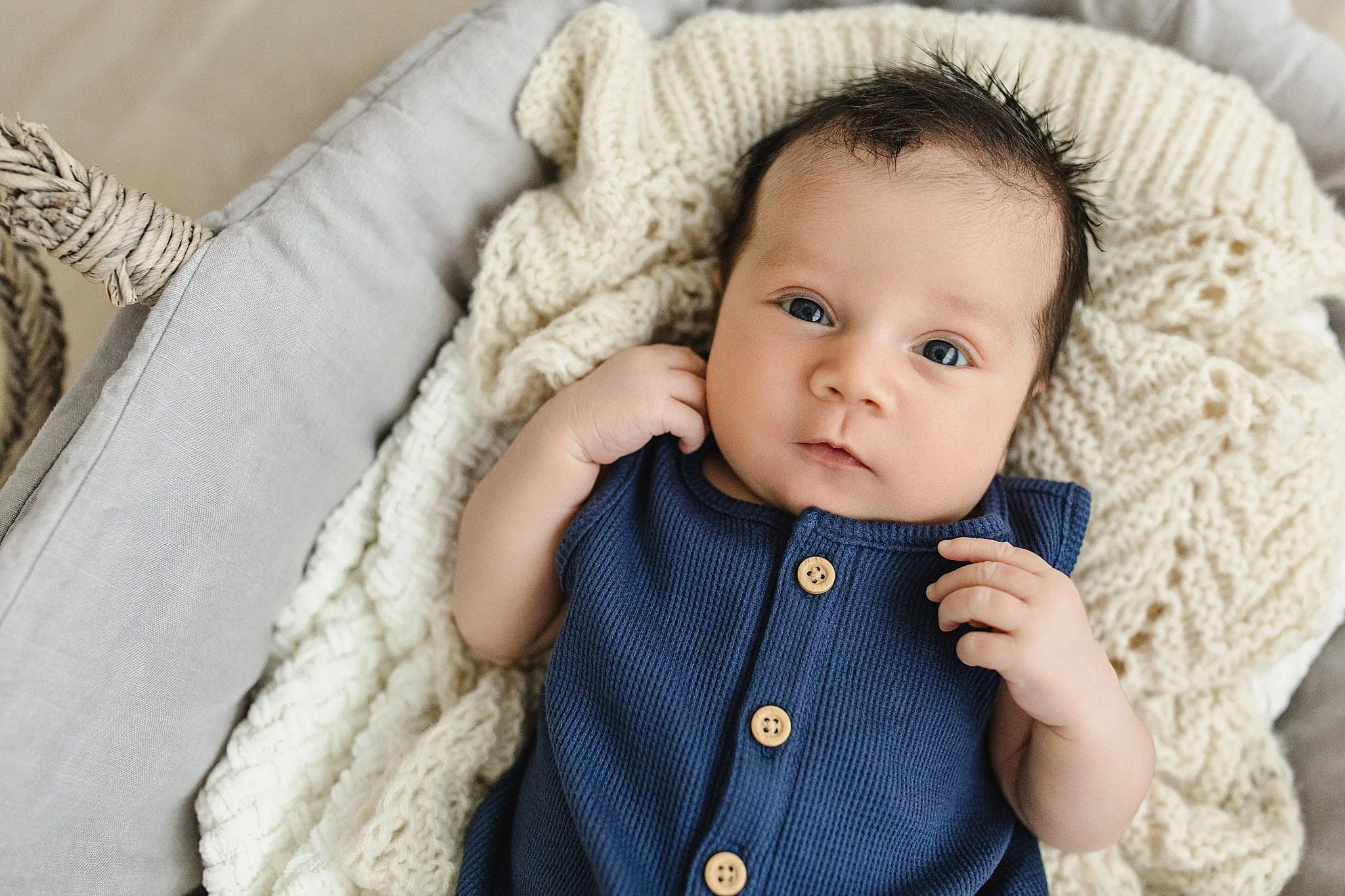 Close-up of a baby lying in a crib, wearing a navy blue shirt with wooden buttons, with a knitted blanket and blanket in the crib during a lifestyle newborn photo session with Ally and B Photography.