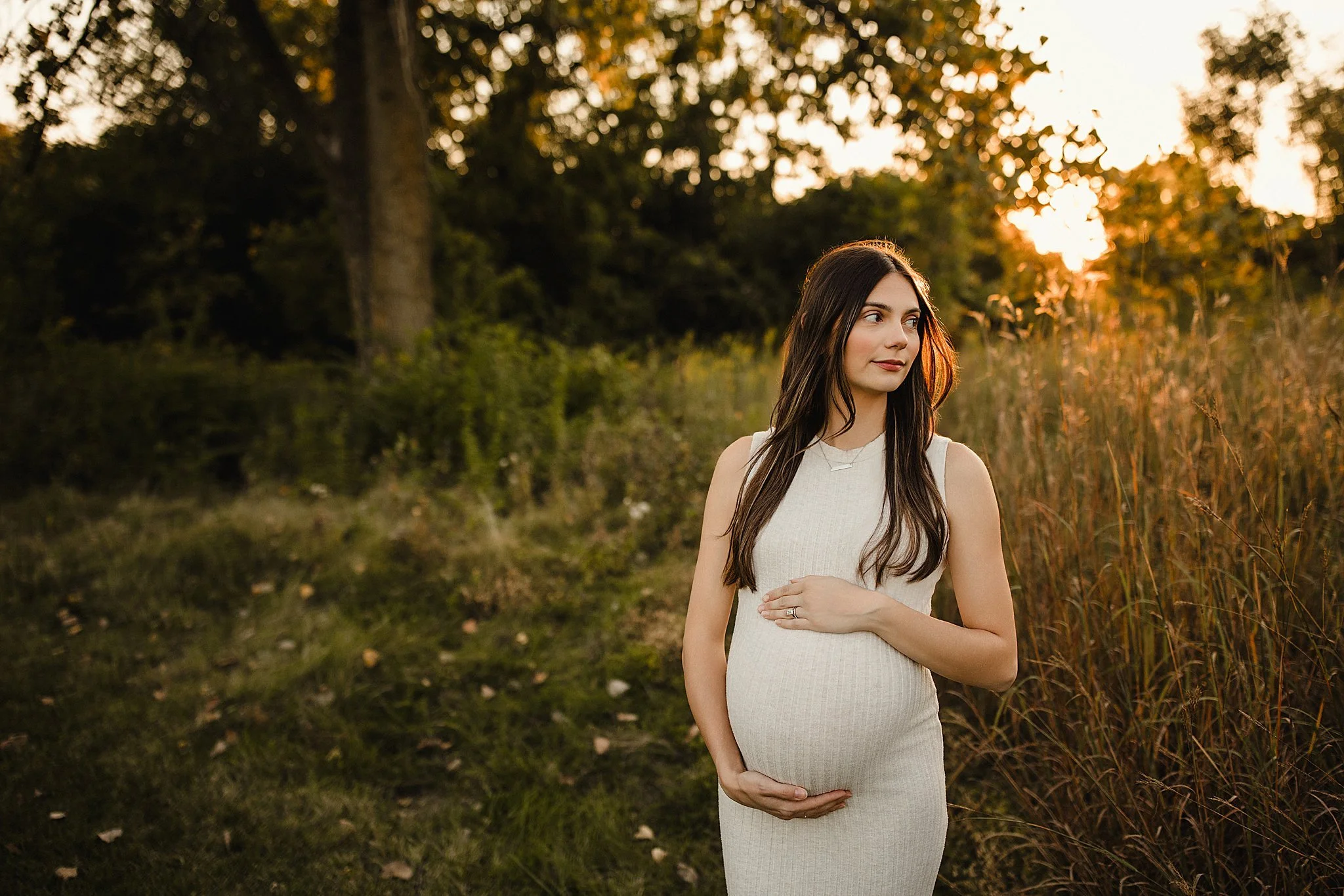 A pregnant woman in a white sleeveless dress standing outdoors during sunset, with greenery and trees in the background with Ally and B Photography for maternity photos outdoor.