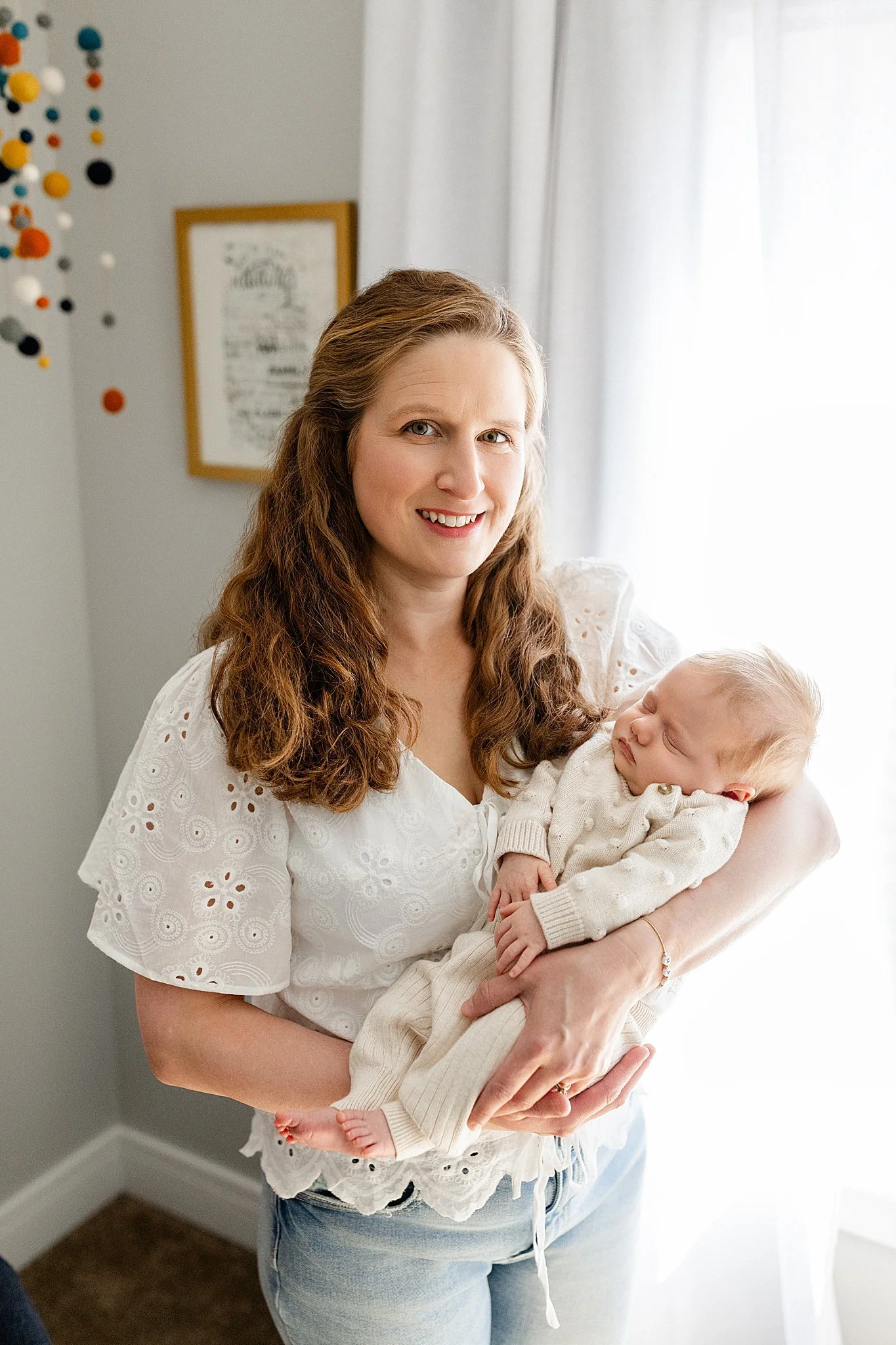 A woman with long, wavy brown hair smiling and holding a baby with light hair. The woman is wearing a white eyelet blouse and jeans. The baby is dressed in a cream-colored sweater and pants with the best family photographers in Naperville, IL.