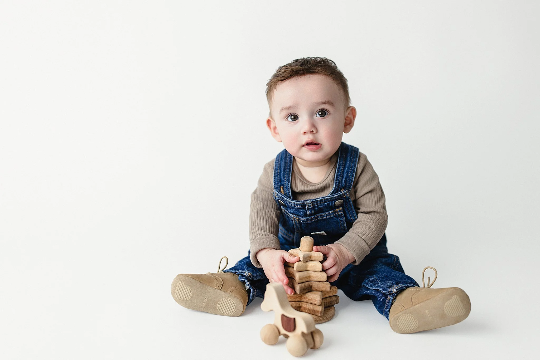 Baby sitting on the floor playing with wooden stacking blocks and a wooden horse toy, wearing denim overalls and tan shoes, against a plain white background.