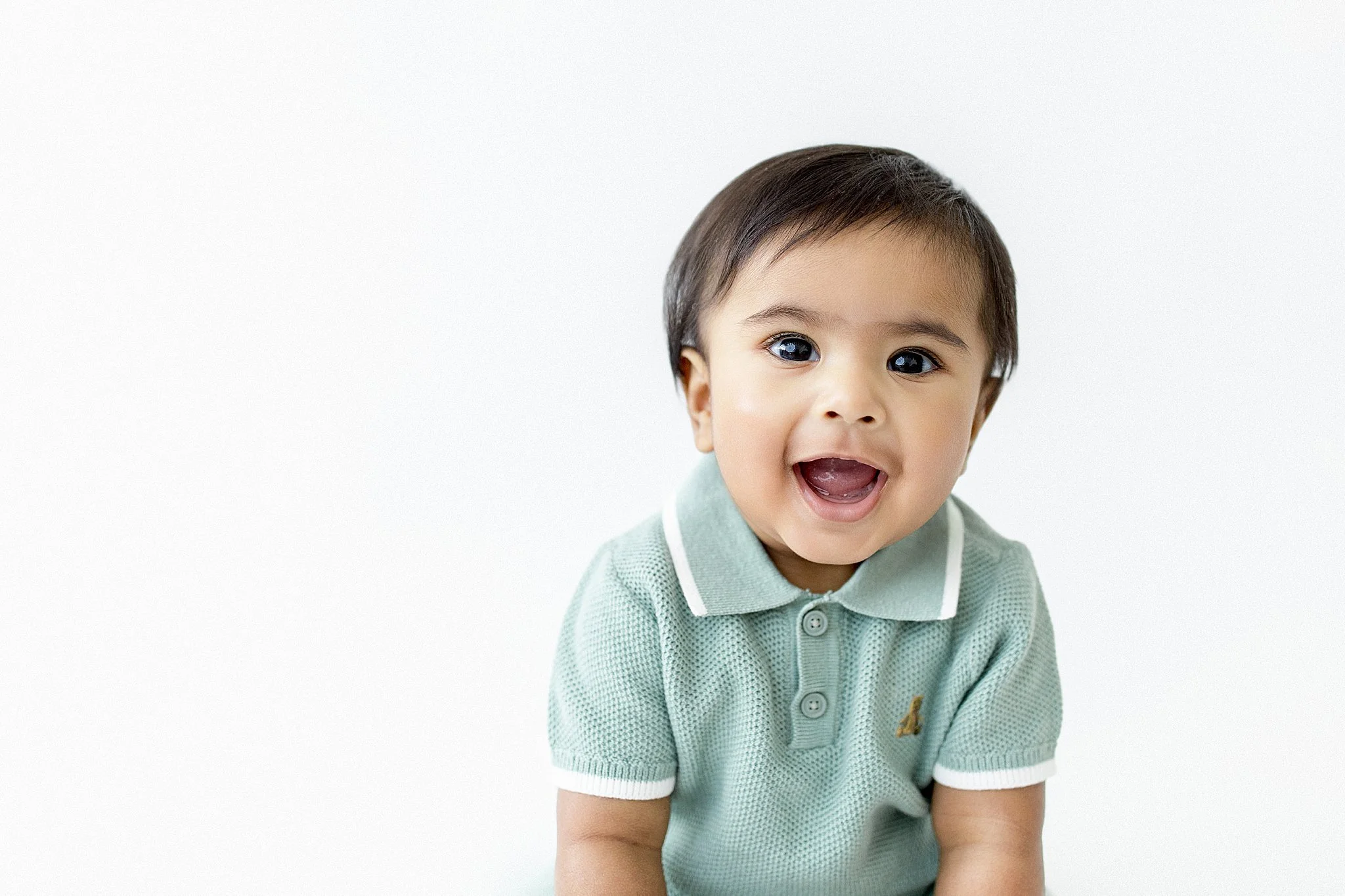 Smiling toddler with dark hair and blue eyes wearing a light green collared shirt against a white background during a milestone photo session near me.