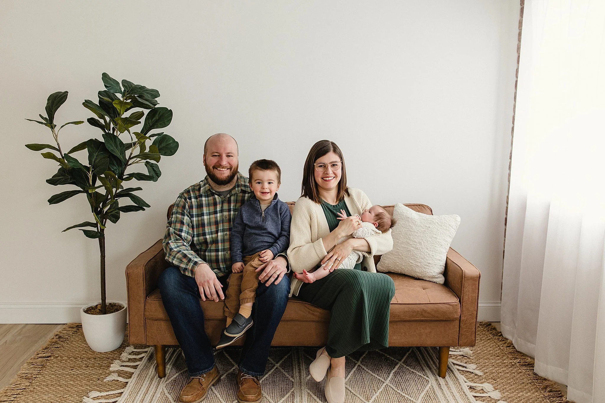 Family of four sitting on a brown sofa in a room with white walls, next to a potted plant and a window with white curtains. The family includes a man, woman, young boy, and a baby girl during lifestyle photography in naperville.