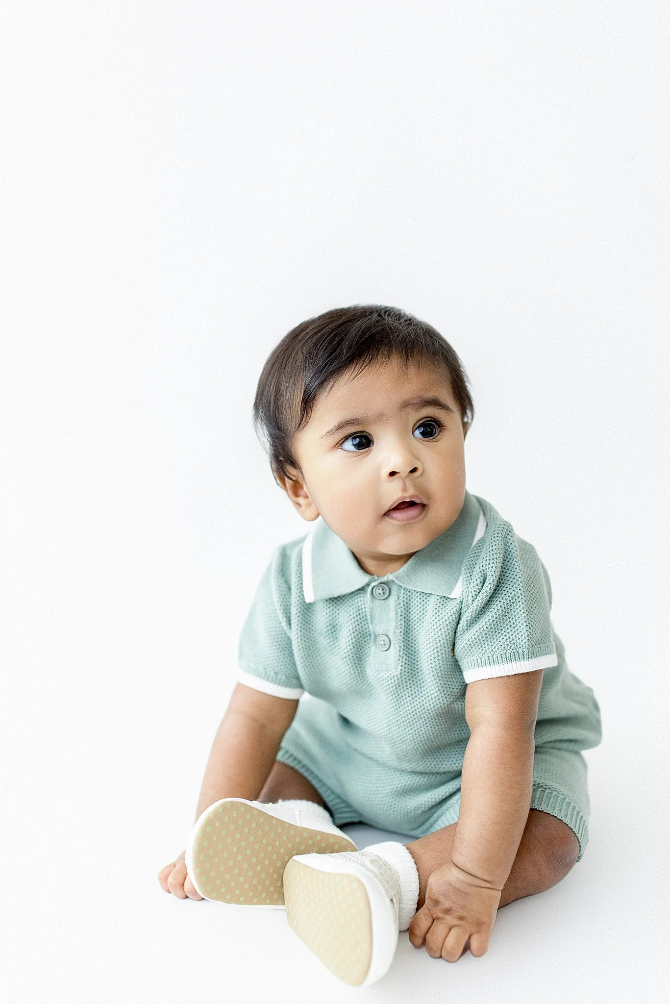 A young child with dark hair, large eyes, and a puzzled expression, sitting on the floor with a mostly white background during a mini milestone session with Ally and B Photography in downtown Naperville.