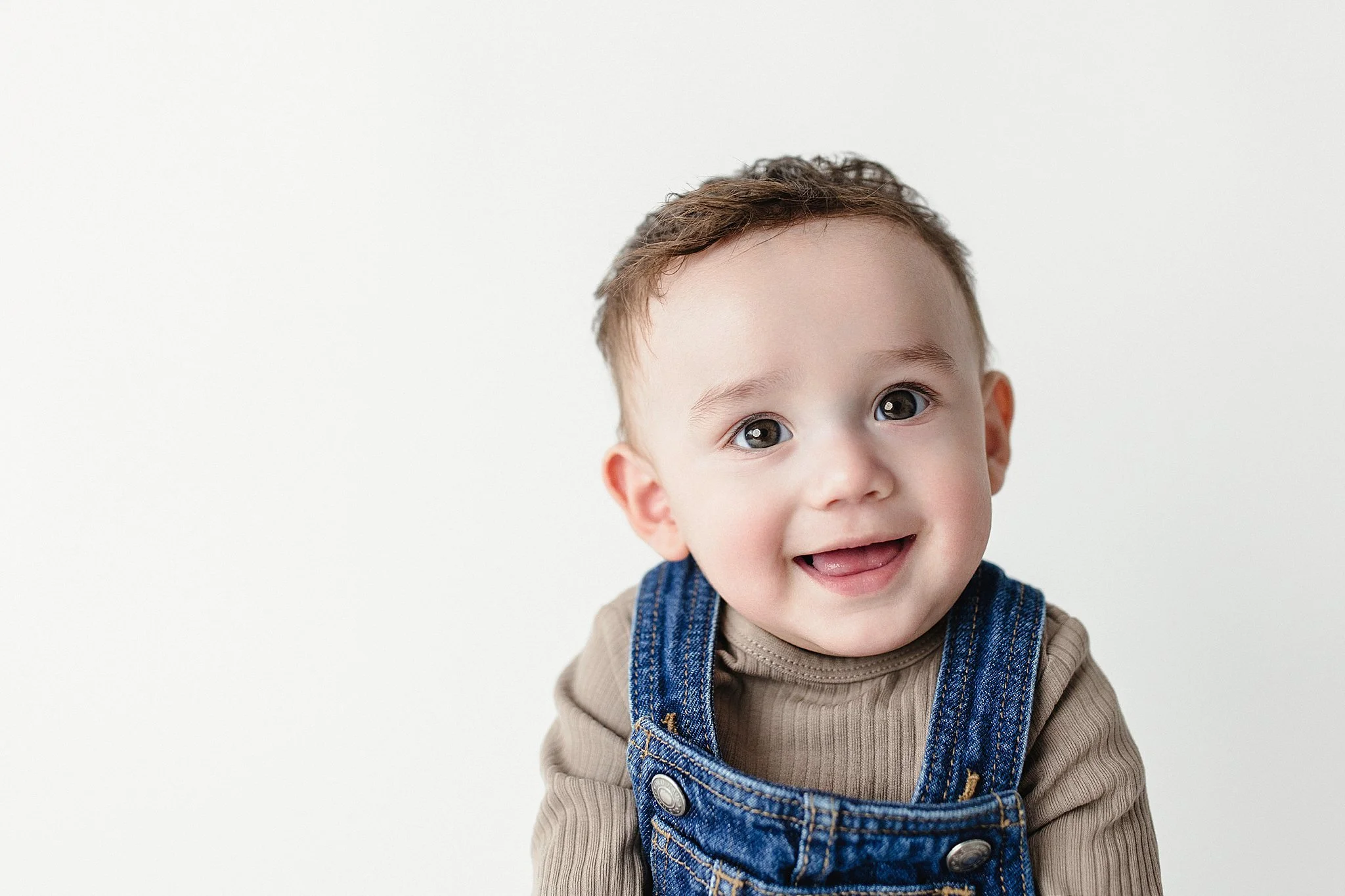 Close-up of smiling toddler boy with brown hair, wearing a beige long-sleeve shirt and denim overalls, against a plain white background.