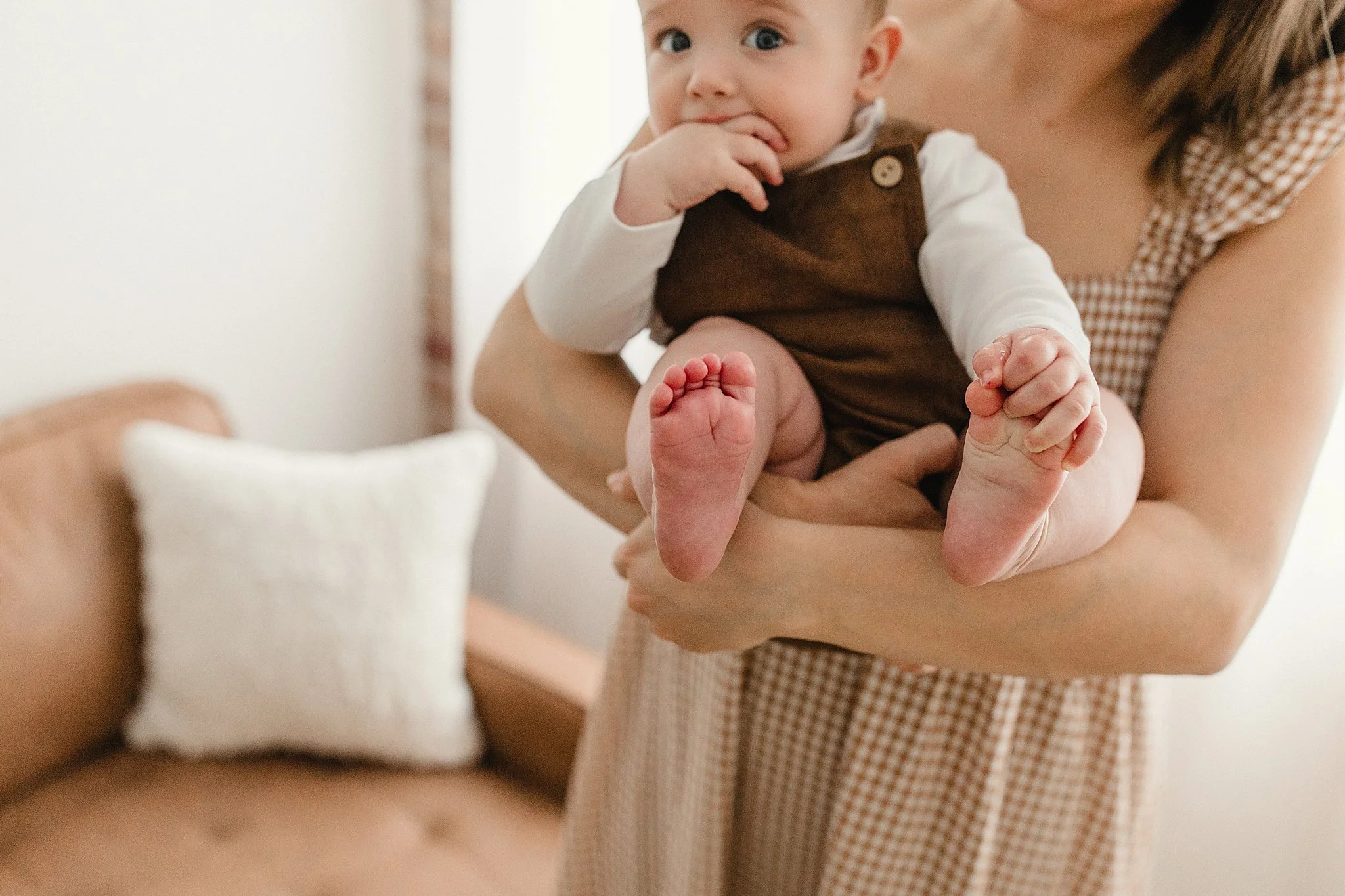 A woman holding a baby inside a room with a brown couch and a white pillow during a family photoshoot near me.
