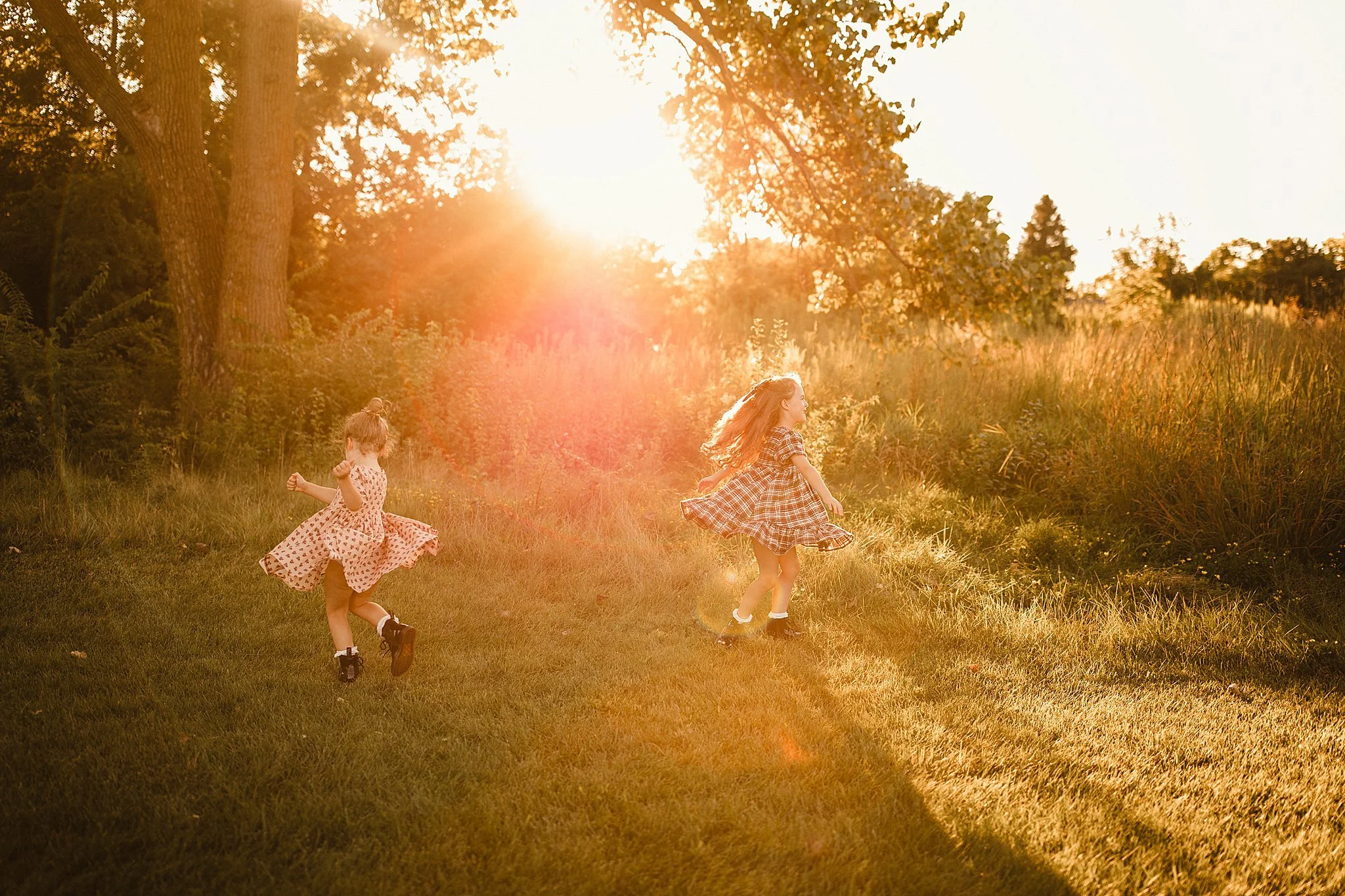 Two young girls in dresses playing and running on a grassy field in a park during sunset with a Naperville family photographer.