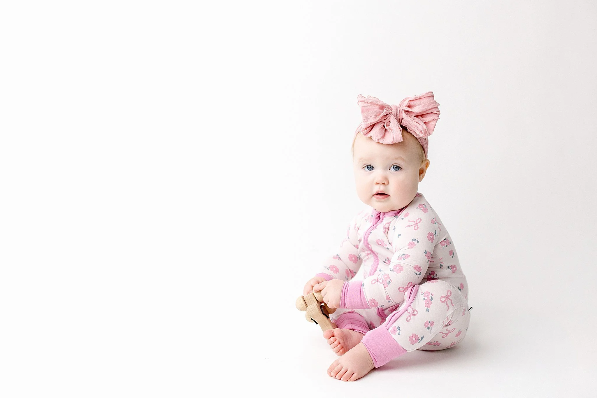A baby girl with blue eyes wearing pink floral pajamas and a large pink bow headband, sitting on a white background, holding a small wooden toy during a simple baby photoshoot with Ally and B Photography.