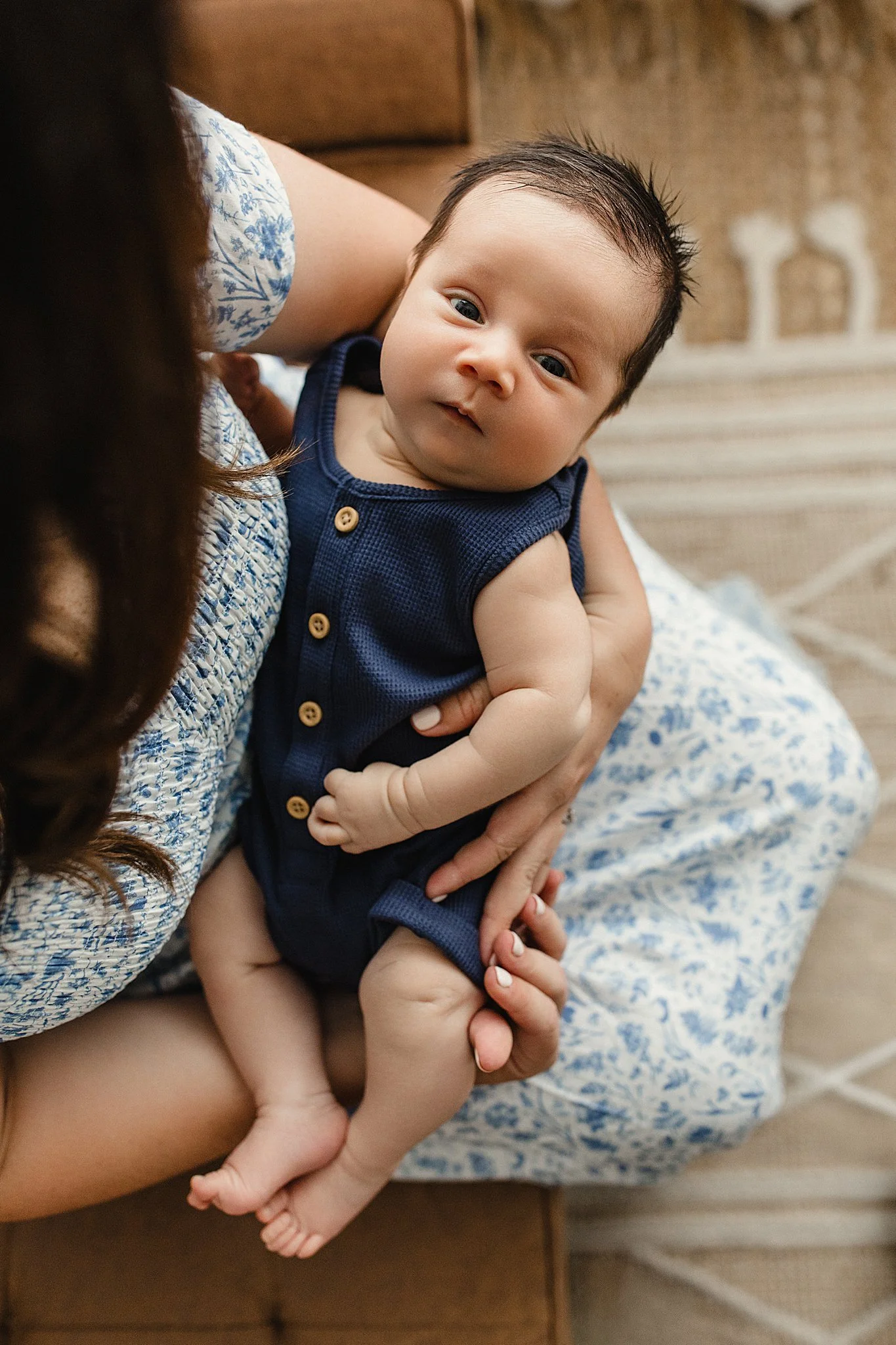 A baby with dark hair and blue eyes being held by a woman, with the woman's arm visible, wearing a floral blue and white dress with newborn photographers in downtown Naperville, IL.
