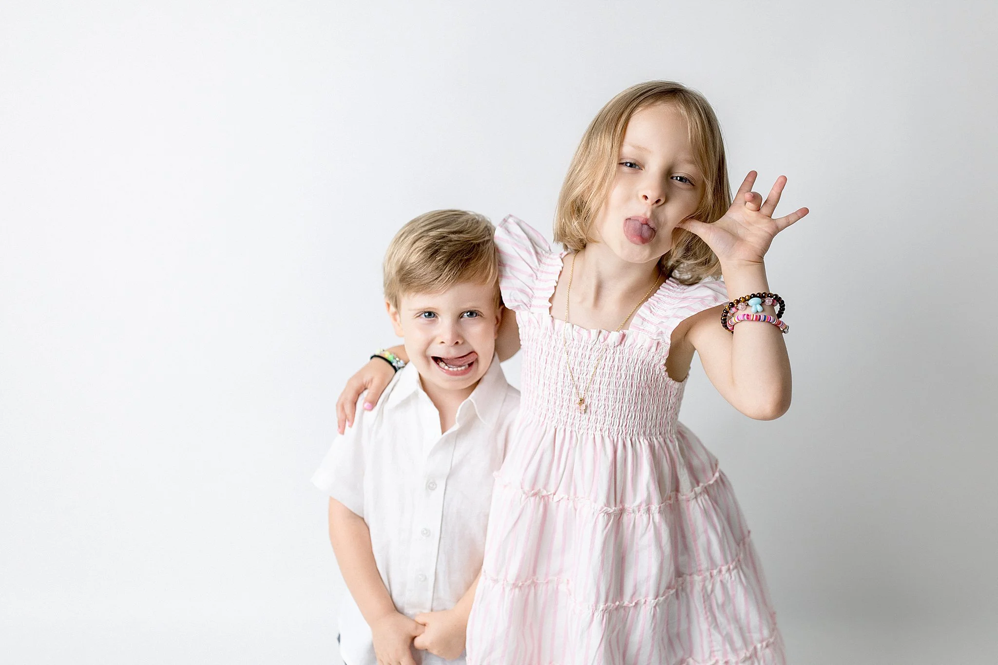 Two children, a girl and a boy, posing playfully against a plain white background. The girl is sticking her tongue out and making a peace sign with her fingers, while the boy is smiling for personality minis near me.