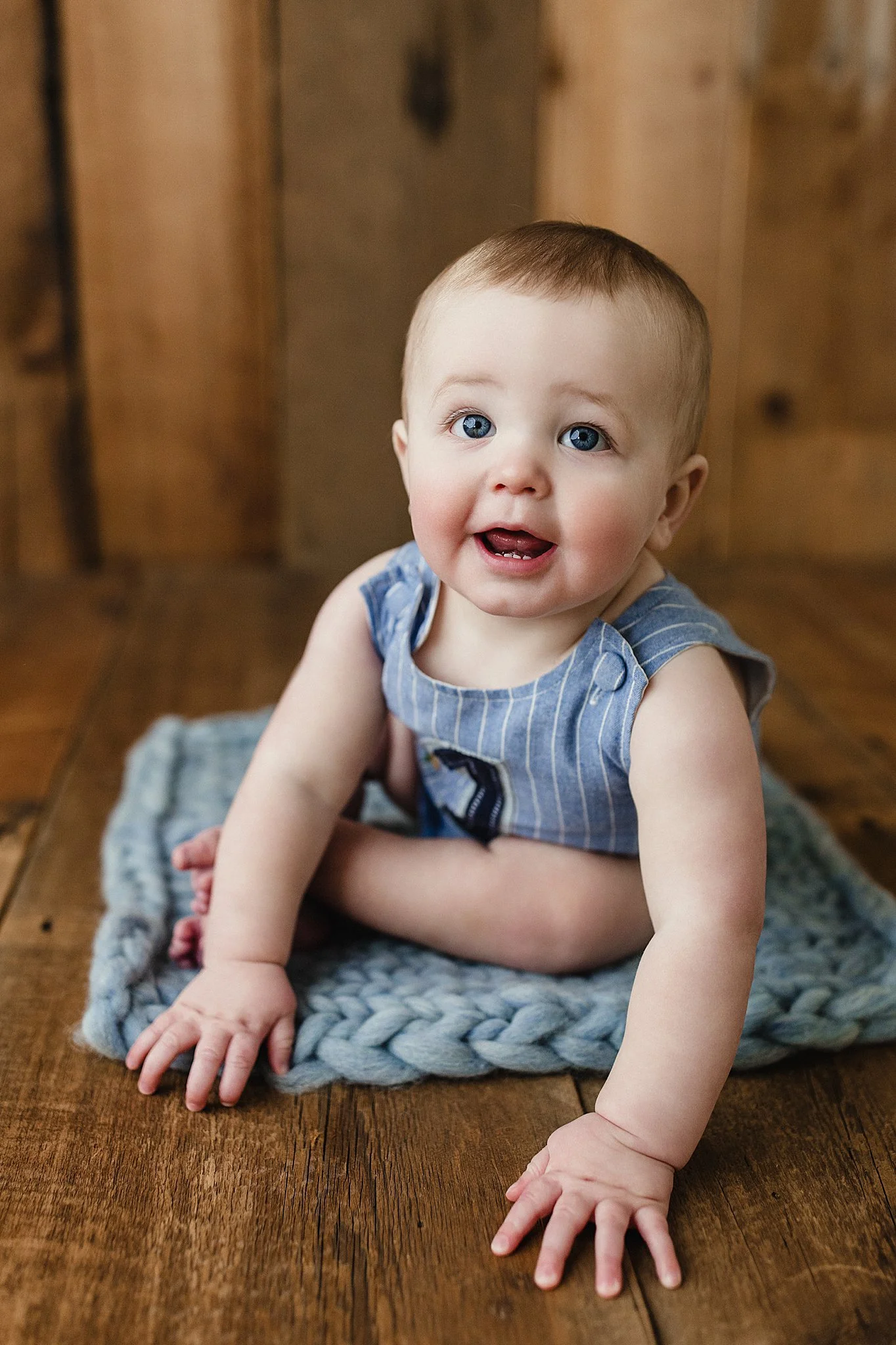 A baby with blue eyes, wearing a blue striped sleeveless shirt, is crawling on a wooden floor on a gray knitted blanket, looking at the camera with a happy expression during a one year old photoshoot with Ally and B Photography.