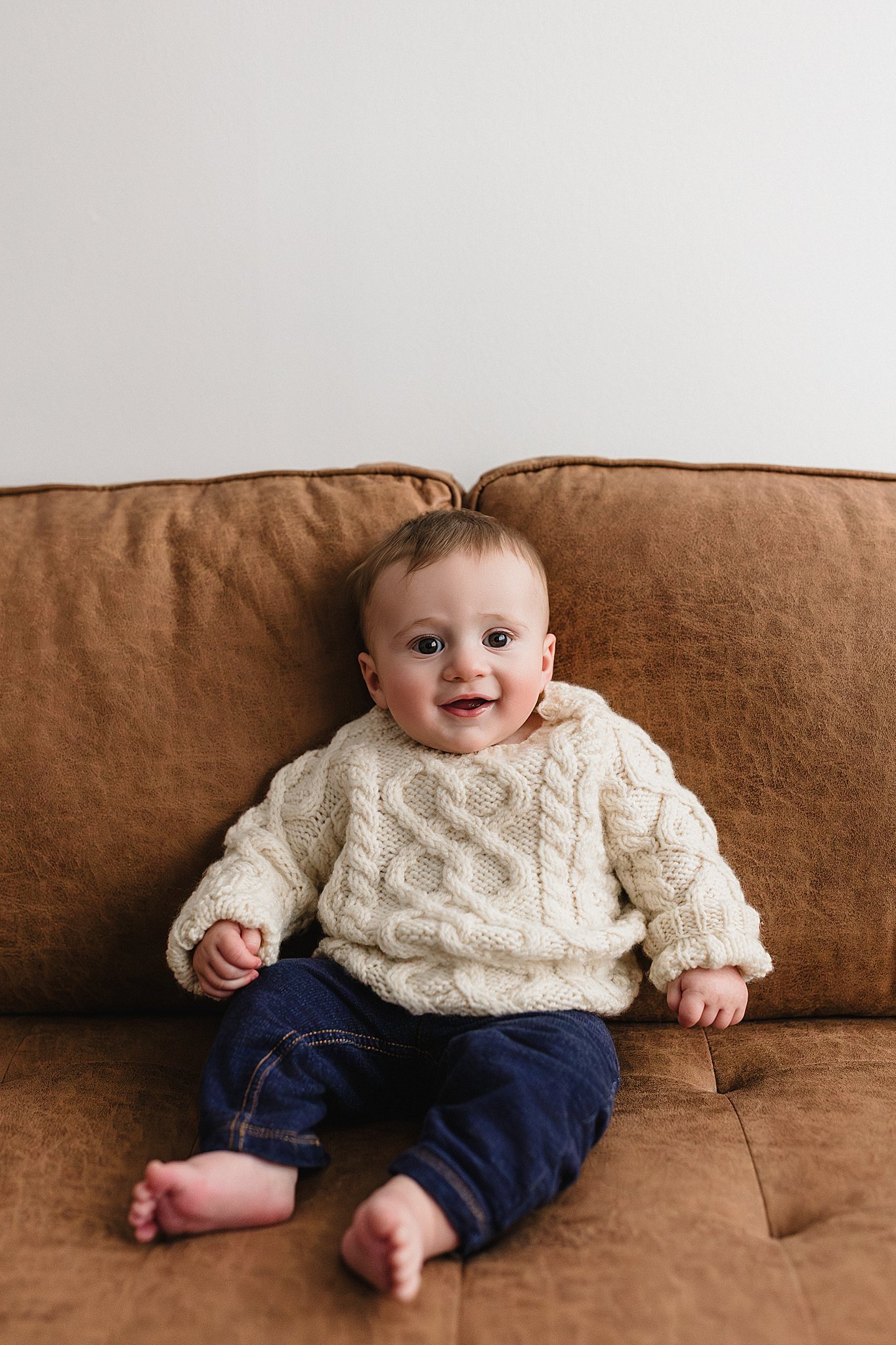 A smiling baby sitting on a brown couch, wearing a cream-colored cable knit sweater and dark denim jeans with a baby photographer near Naperville, IL.