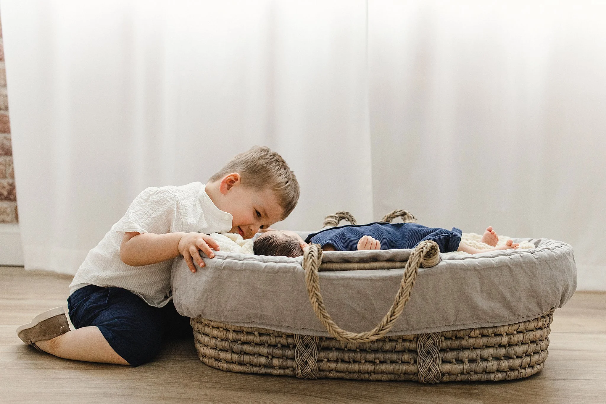 A young boy kneeling on the floor and looking into a woven basket with a baby lying inside, in a room with plain white walls during newborn photography near me.