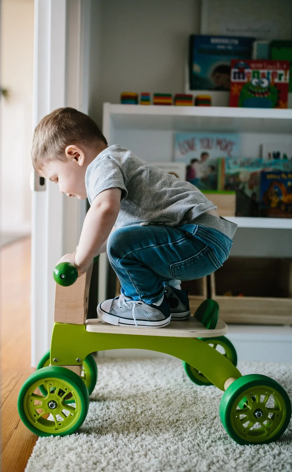Toddler on a bicycle inside with an organized kids bookshelf.