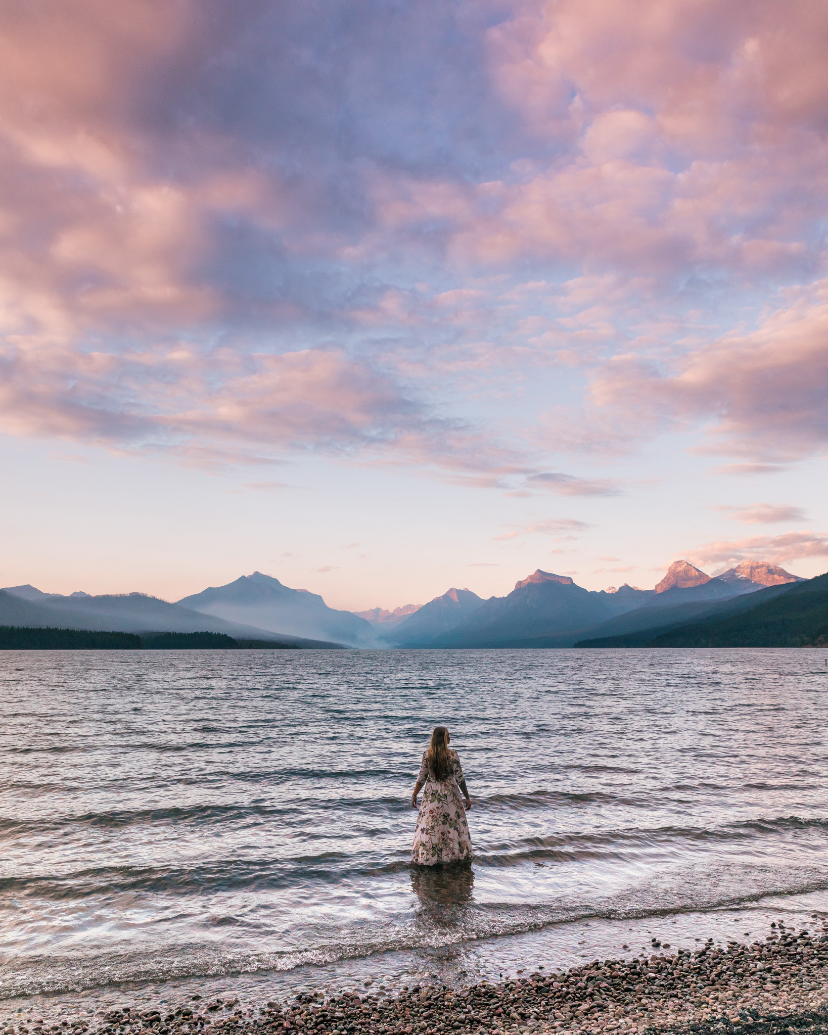 Girl in flowy dress looks at the pink sunset at Lake McDonald in Glacier National Park Montana