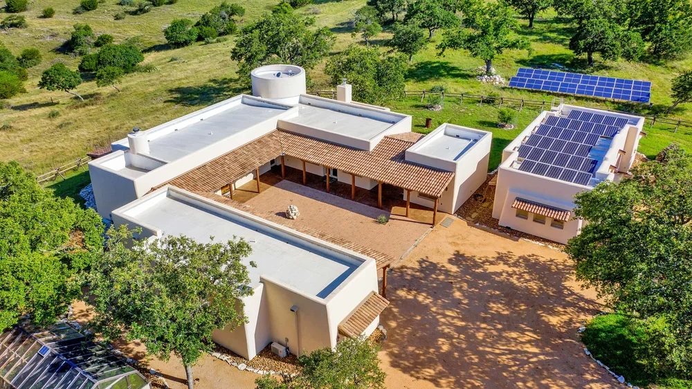 Aerial view of a residential home featuring solar panels on the roof, surrounded by greenery and a clear blue sky.
