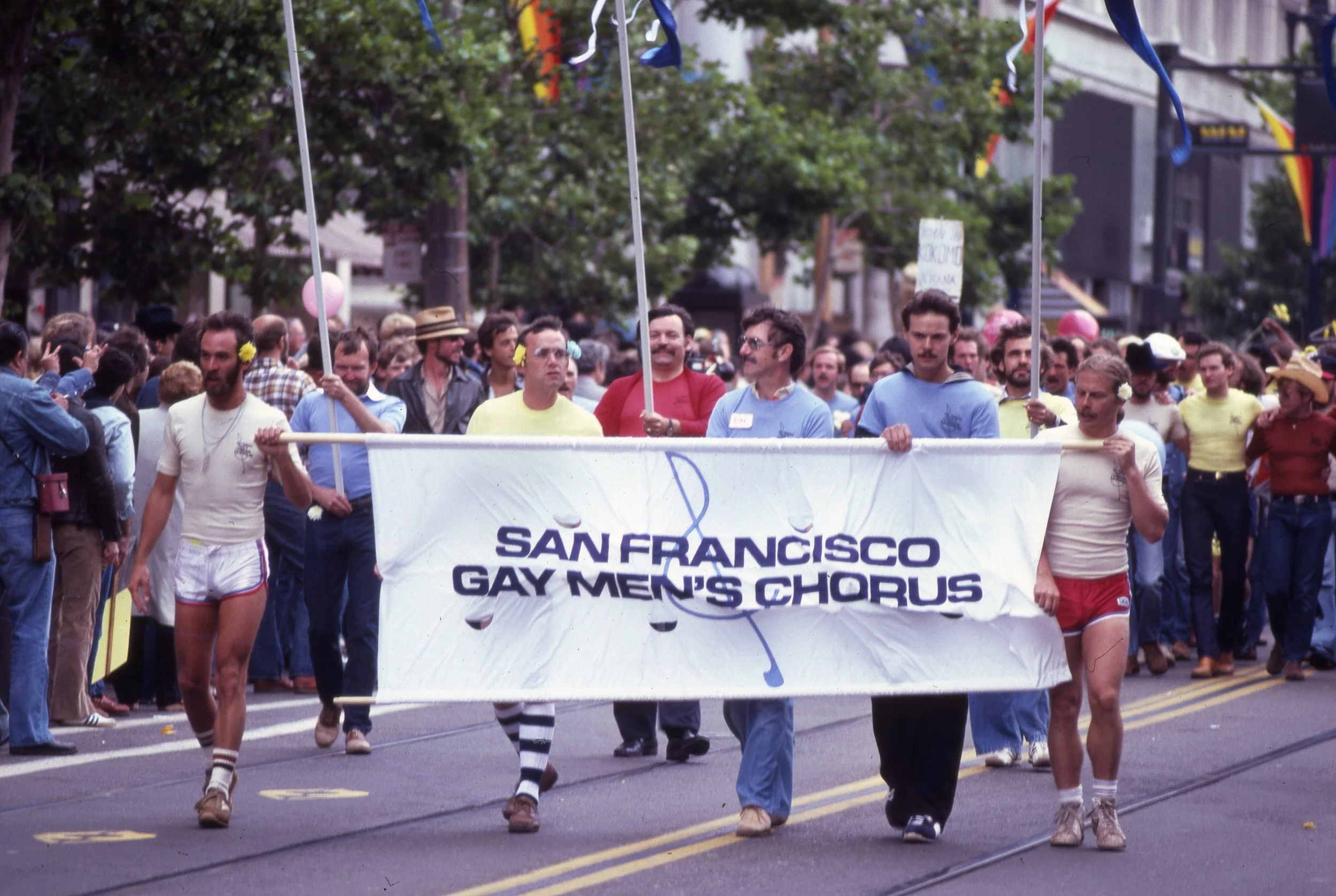 Members of the San Francisco Gay Men’s Chorus marching in a Pride parade carrying an SFGMC banner.