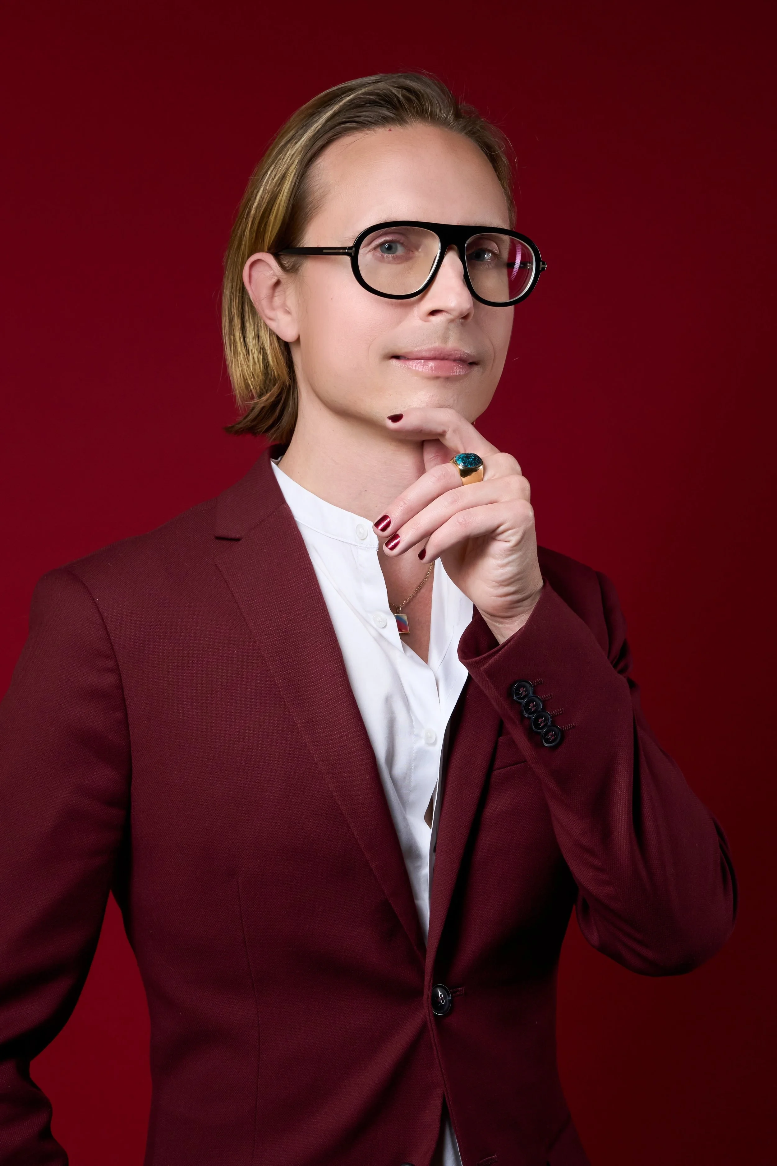 A photograph of Lane Sugata wearing glasses, with a ringed hand under their chin