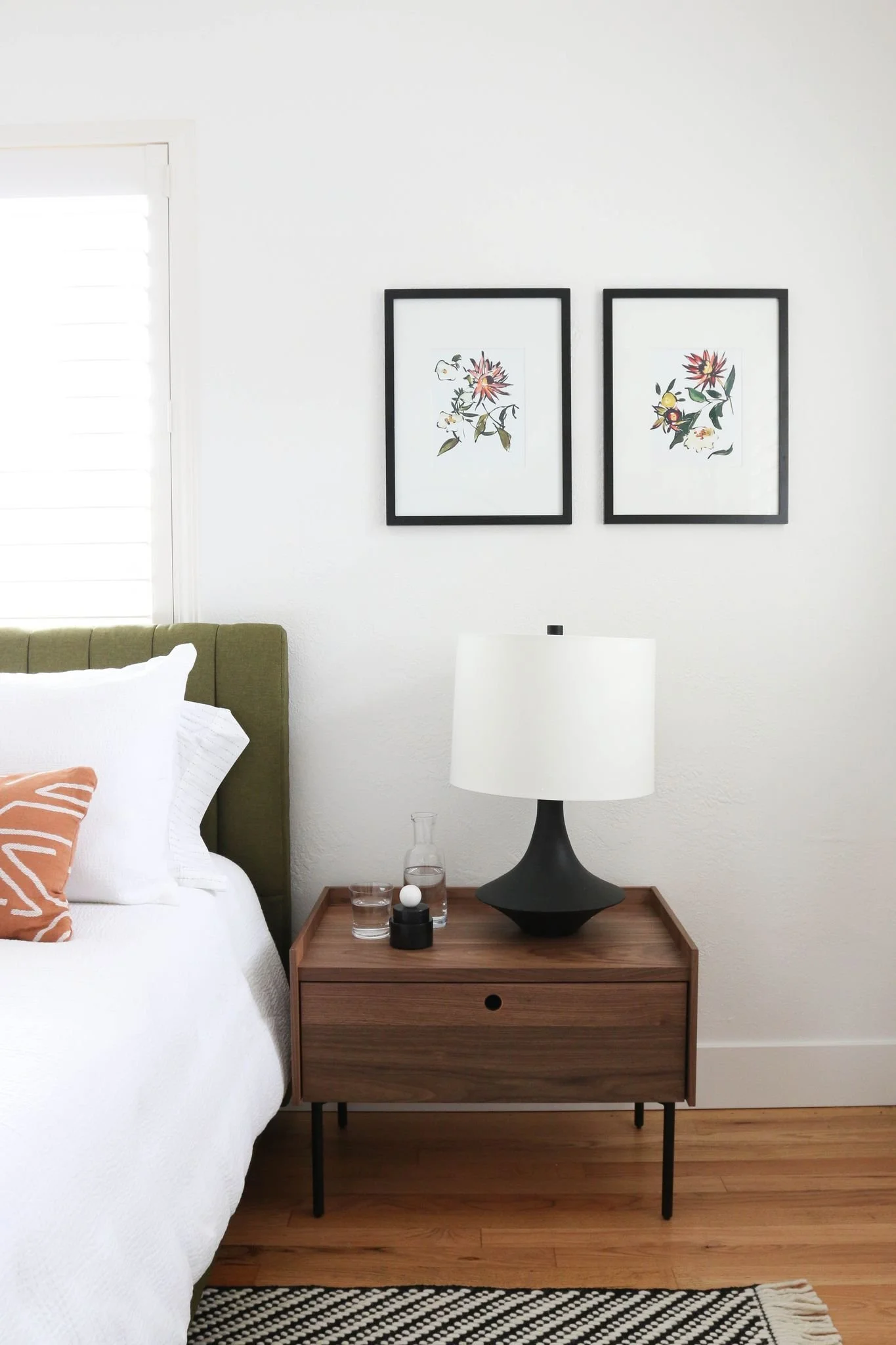 Bedroom with a mid-century modern style, featuring a wooden nightstand with a black and white lamp, beside a bed with white bedding and an orange patterned pillow. Two framed floral artworks hang on the white wall.