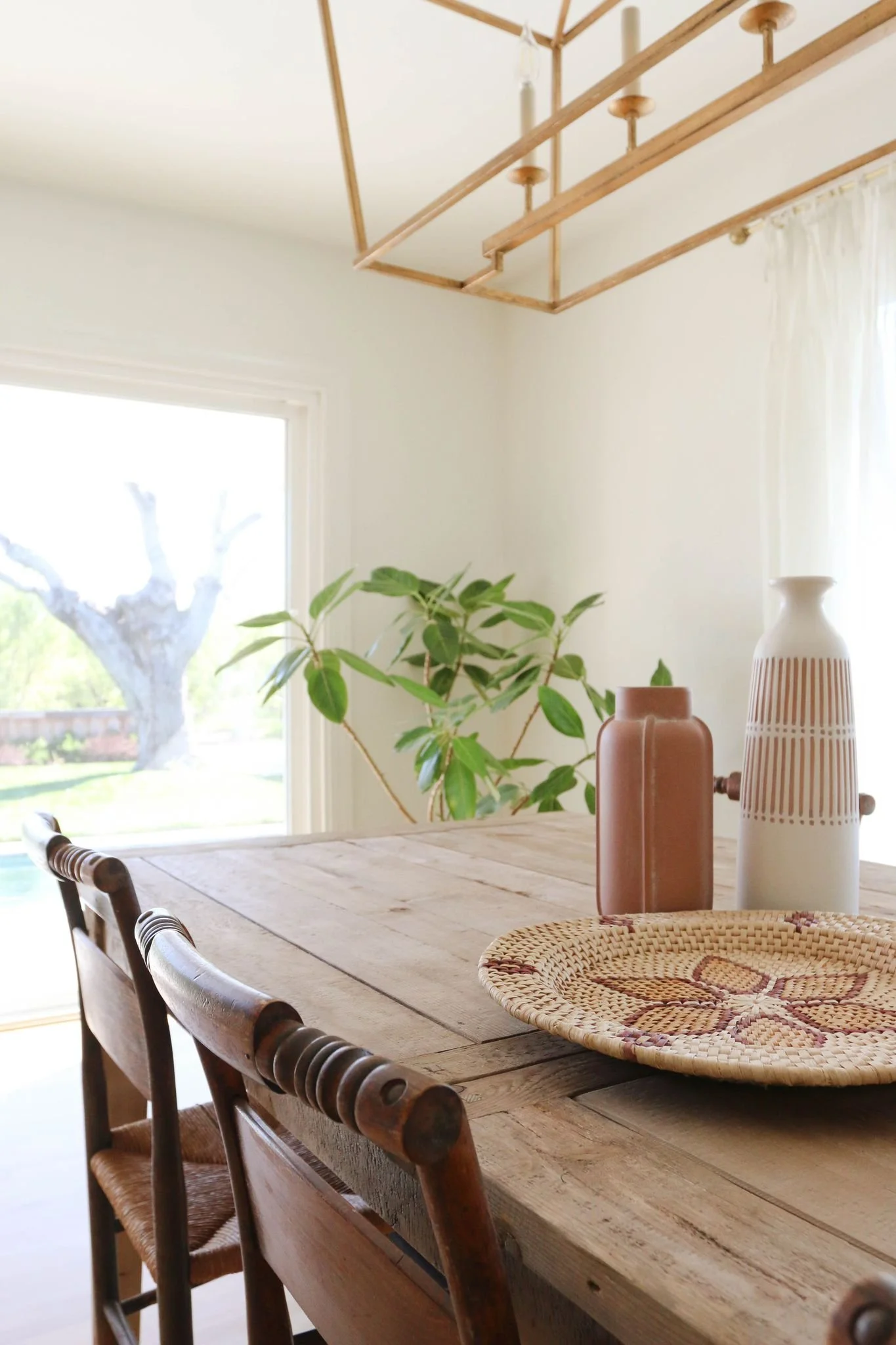 Rustic dining room with wooden table and chairs, decorative vases, woven placemat, large plant, and window view.