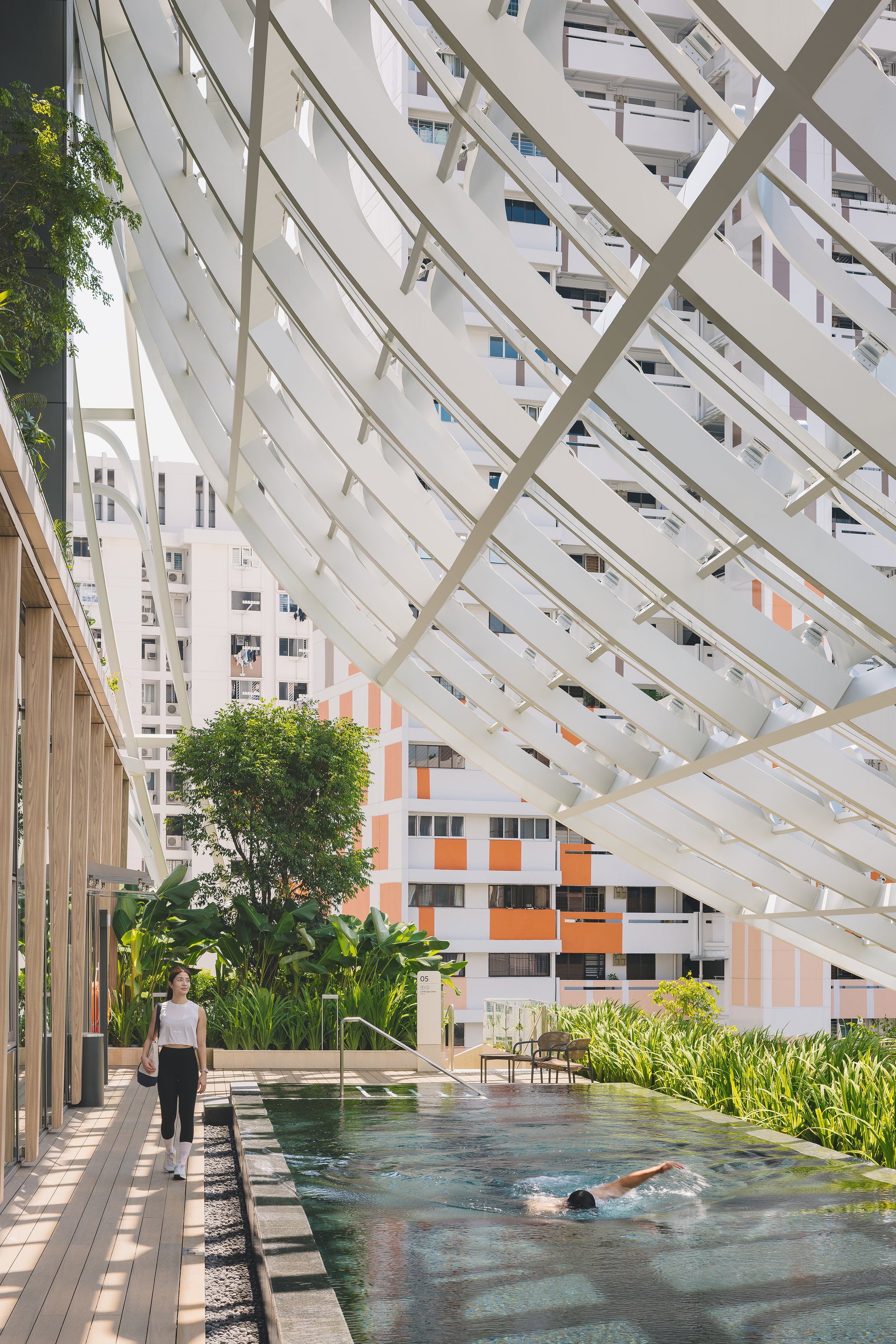 Outdoor lap pool at Keppel South Central beneath a striking white architectural canopy, with lush landscaping, a swimmer in the pool, and a pedestrian walking alongside.