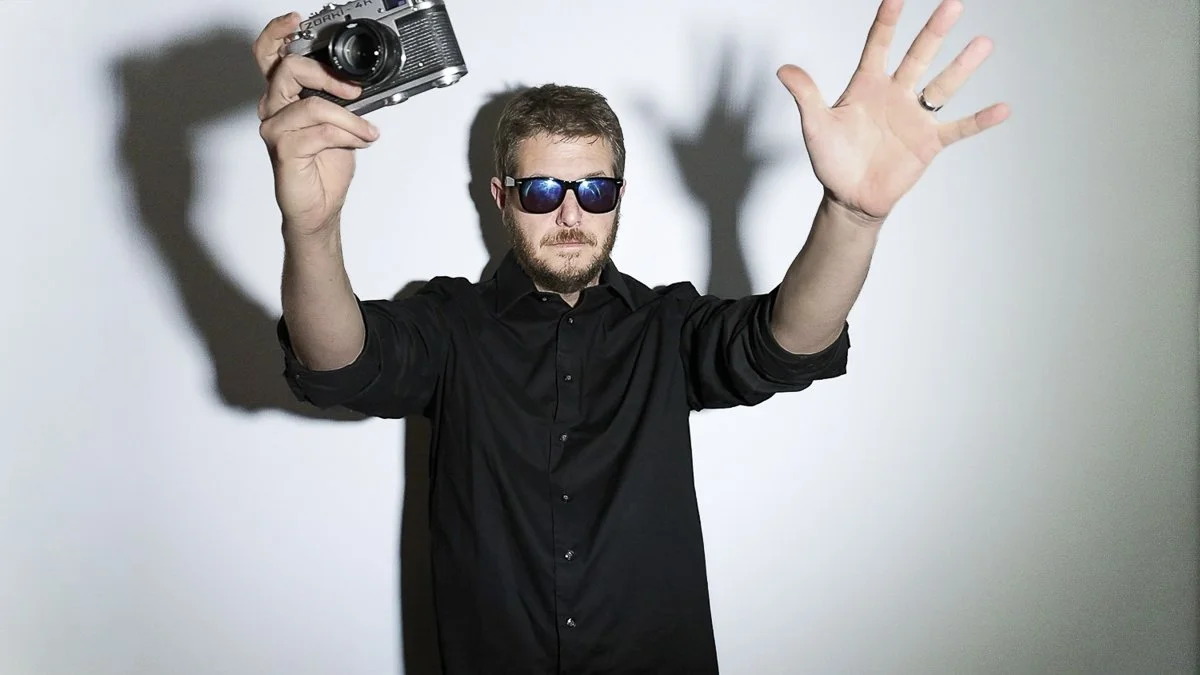 A man wearing sunglasses and a black shirt holding a vintage camera, with his left hand raised in front of a plain background.